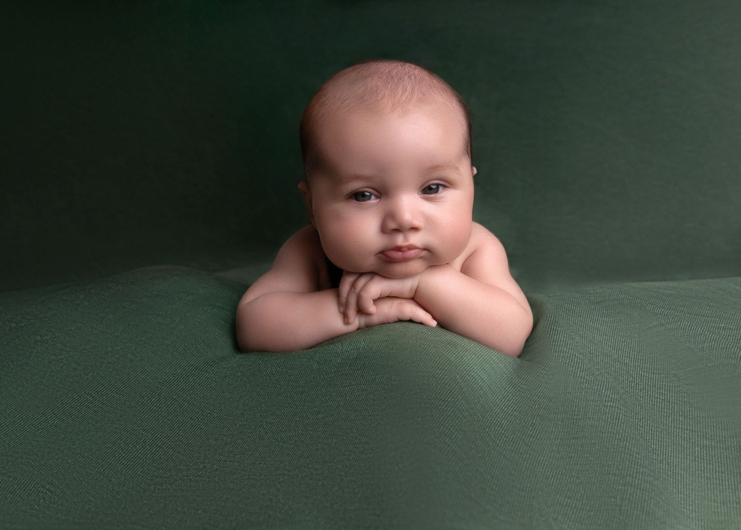 Baby propped on arms against dark green backdrop in studio portrait setting.