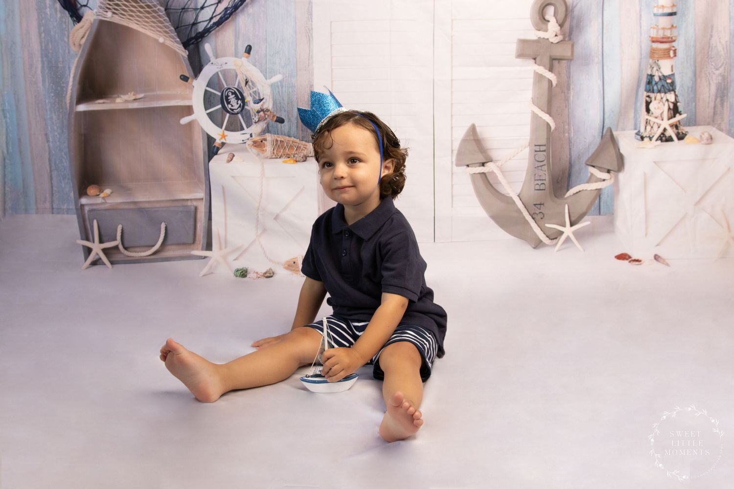 Small child in navy outfit sitting on nautical-themed studio set with lighthouse props.