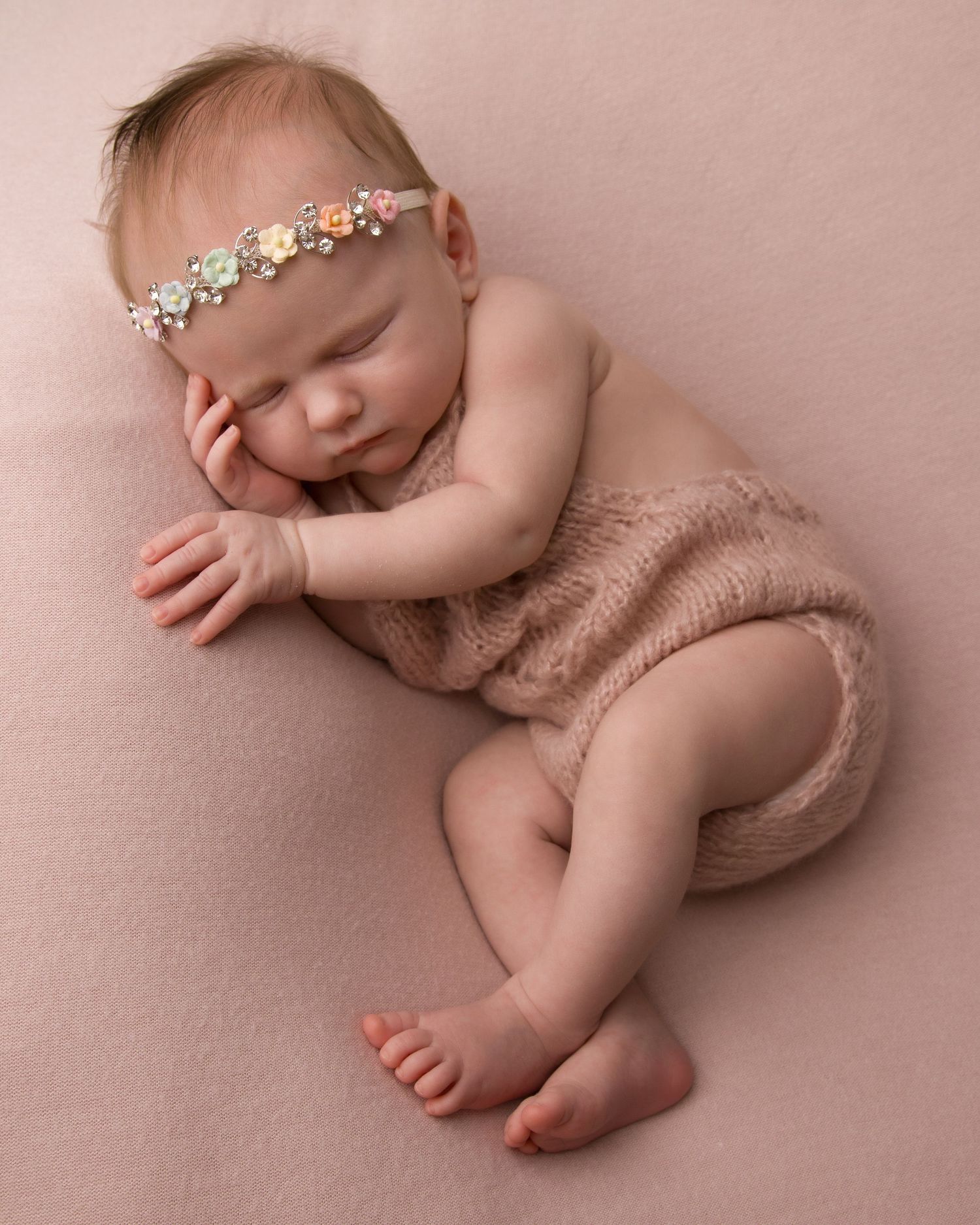 Sleeping newborn wearing floral headband and knit romper on pink backdrop.