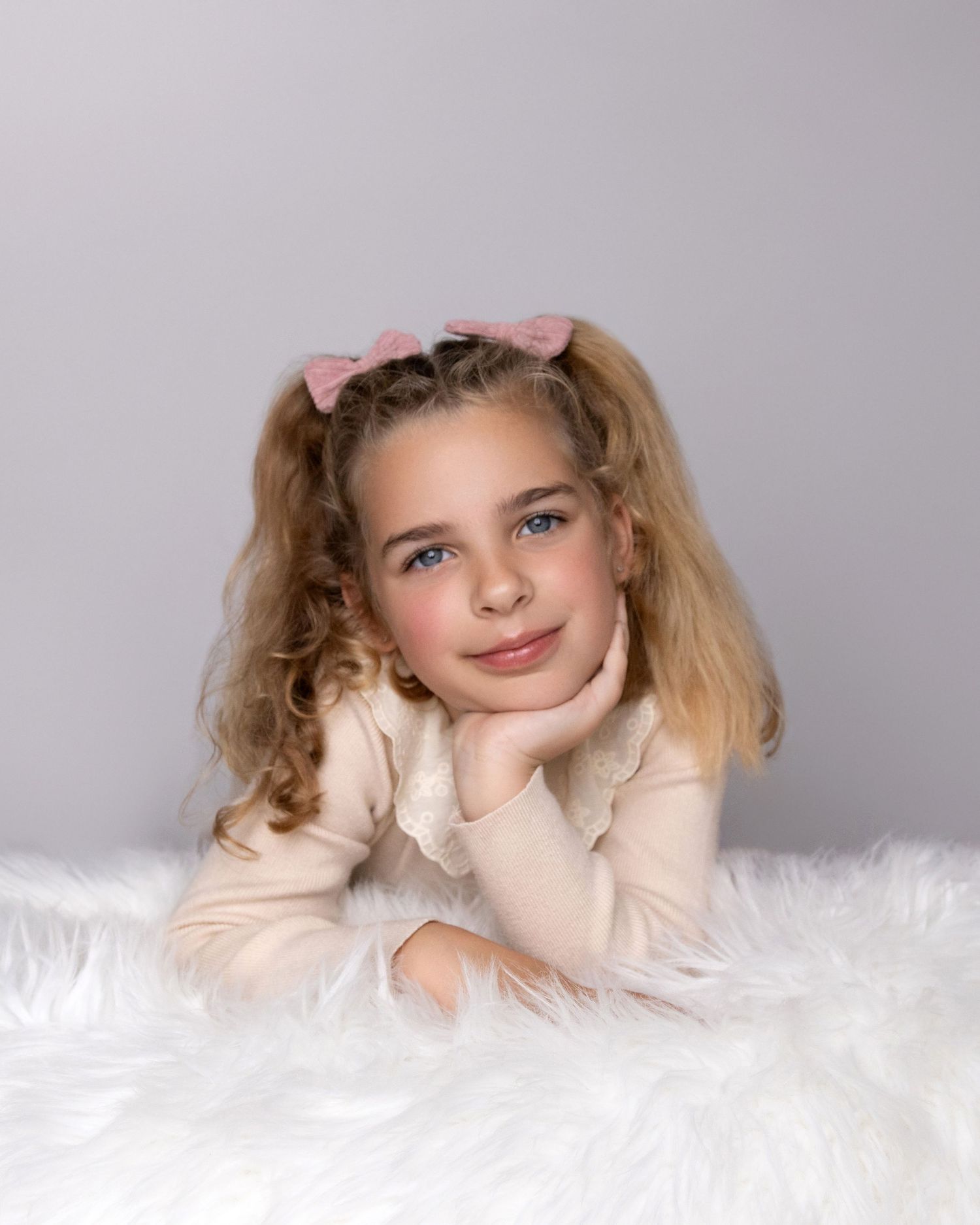 Child in cream sweater lying on white fur backdrop with pink hair bows.