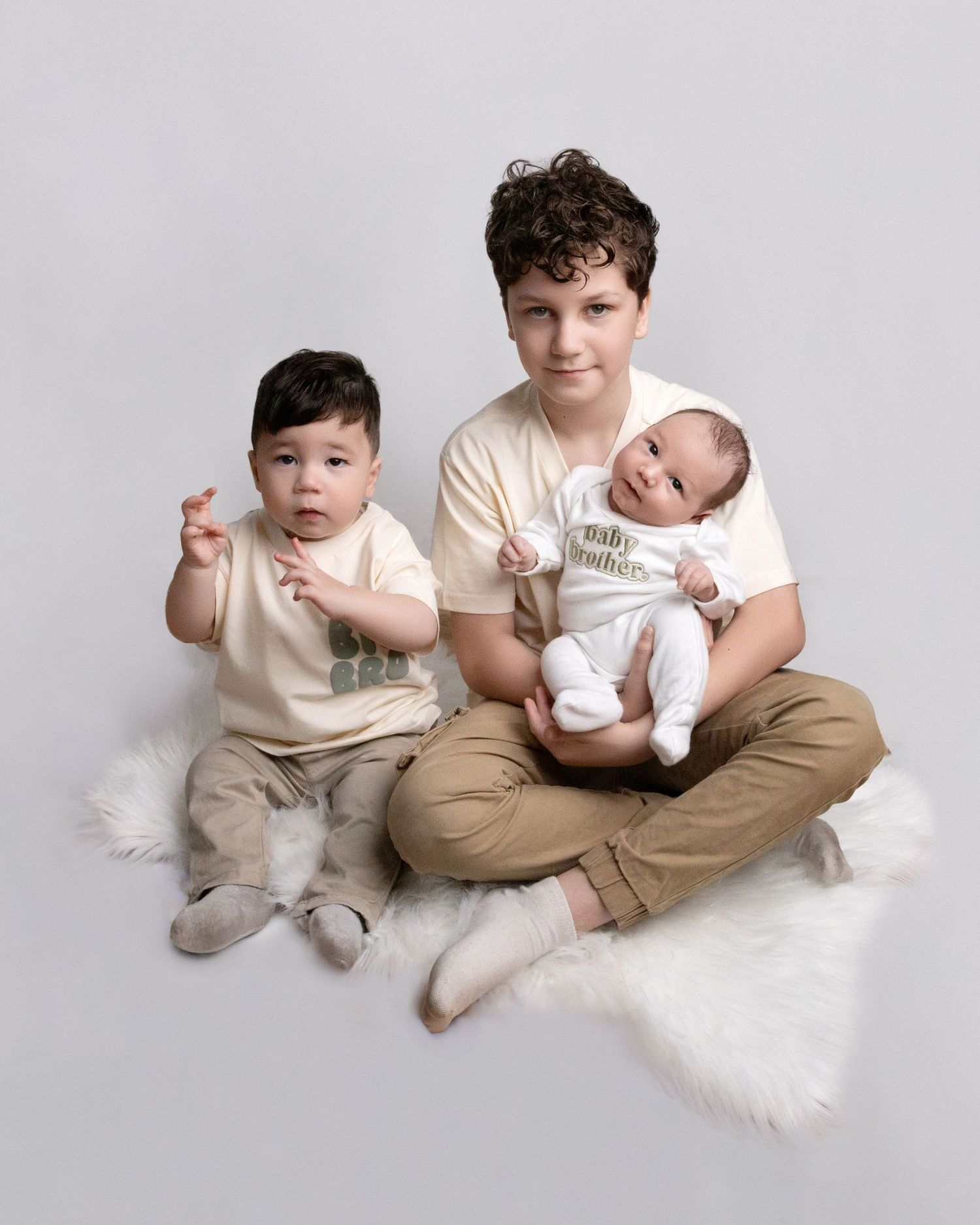 Family portrait on white fur rug with three people in neutral tones against light background.