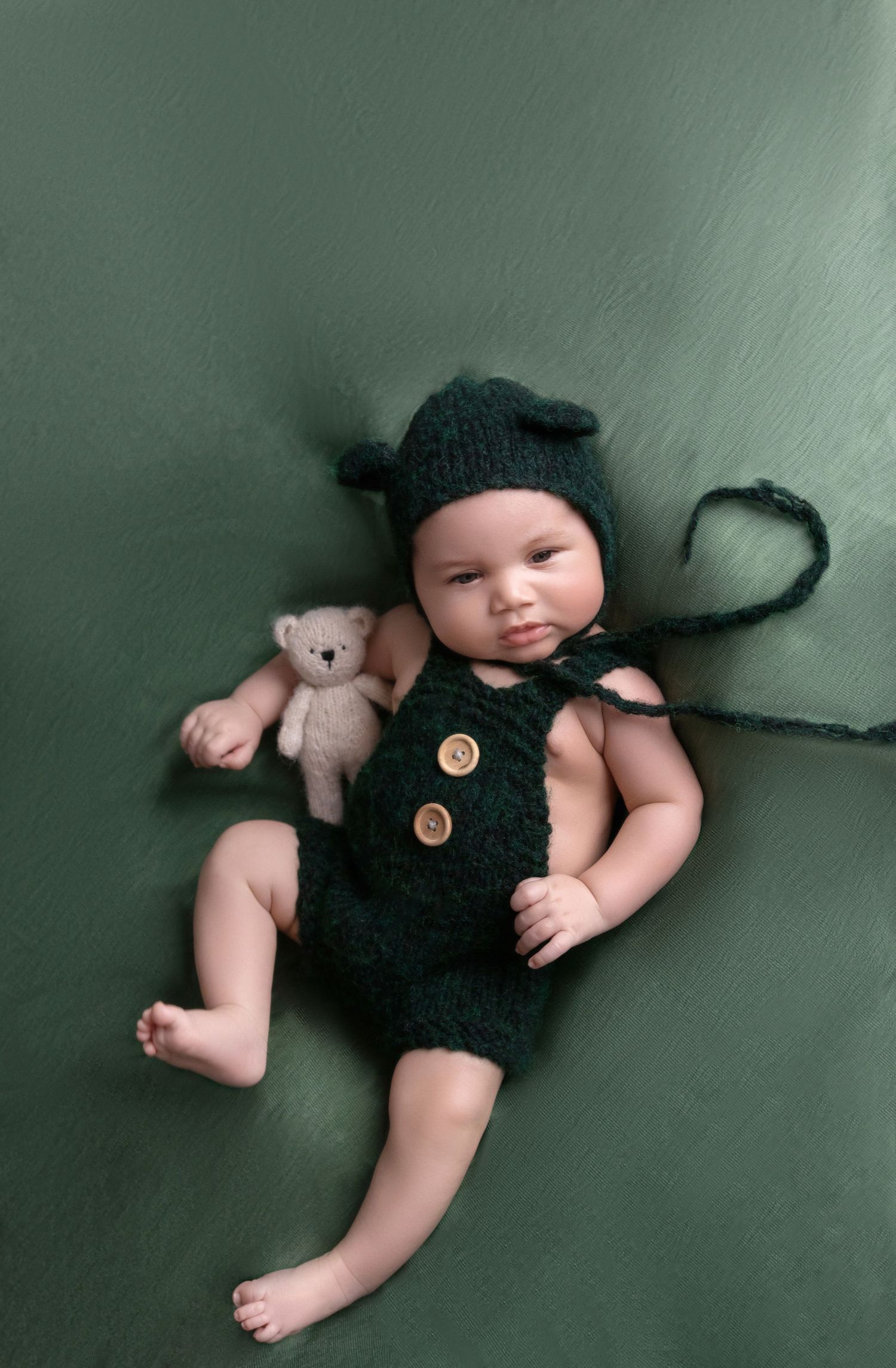 Baby wearing knitted black bear outfit with matching hat lying beside small teddy bear.