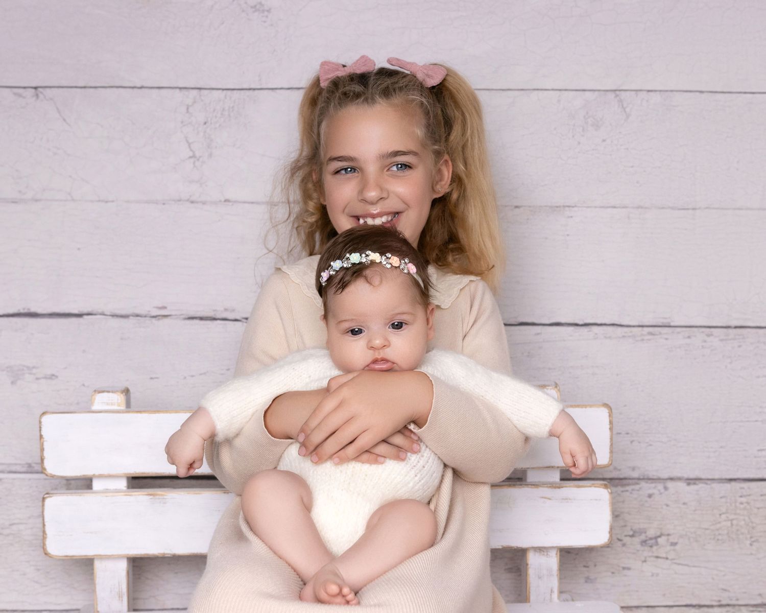 Studio portrait on white bench with young child holding infant in matching white outfits.