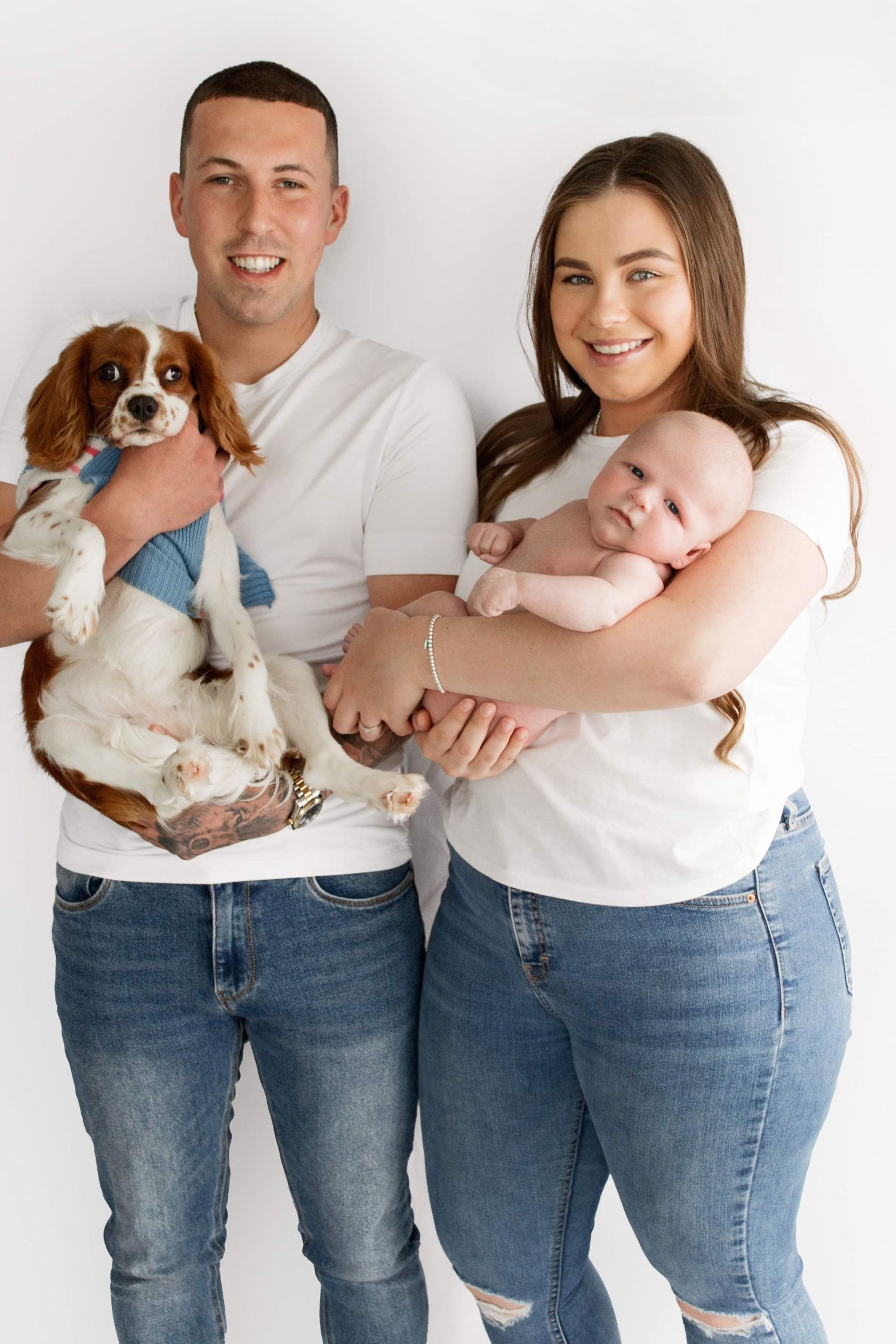 A casual family portrait with a Cavalier King Charles Spaniel puppy and infant against a white background.