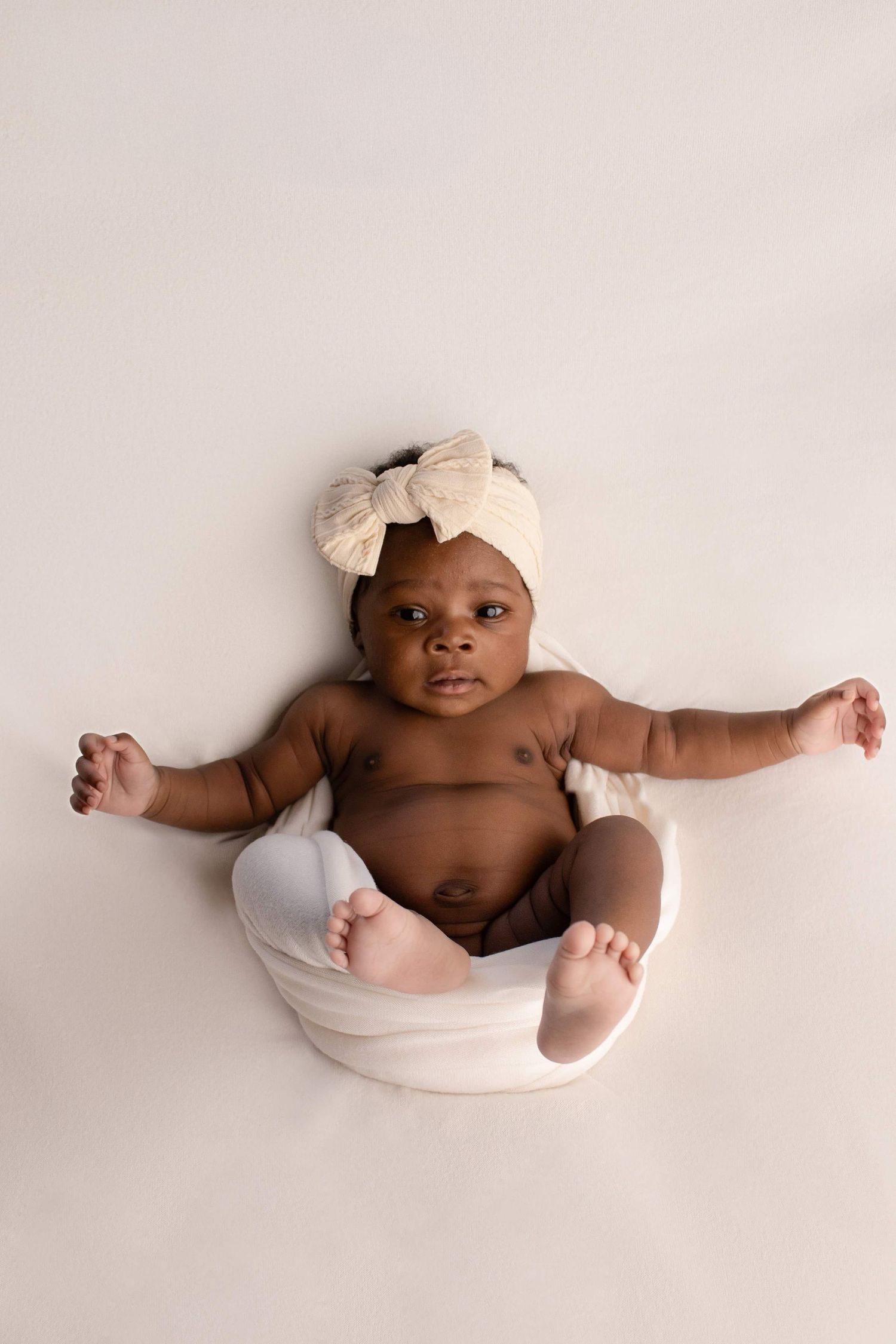 Baby wearing cream bow headwrap poses in white bowl during professional photoshoot.