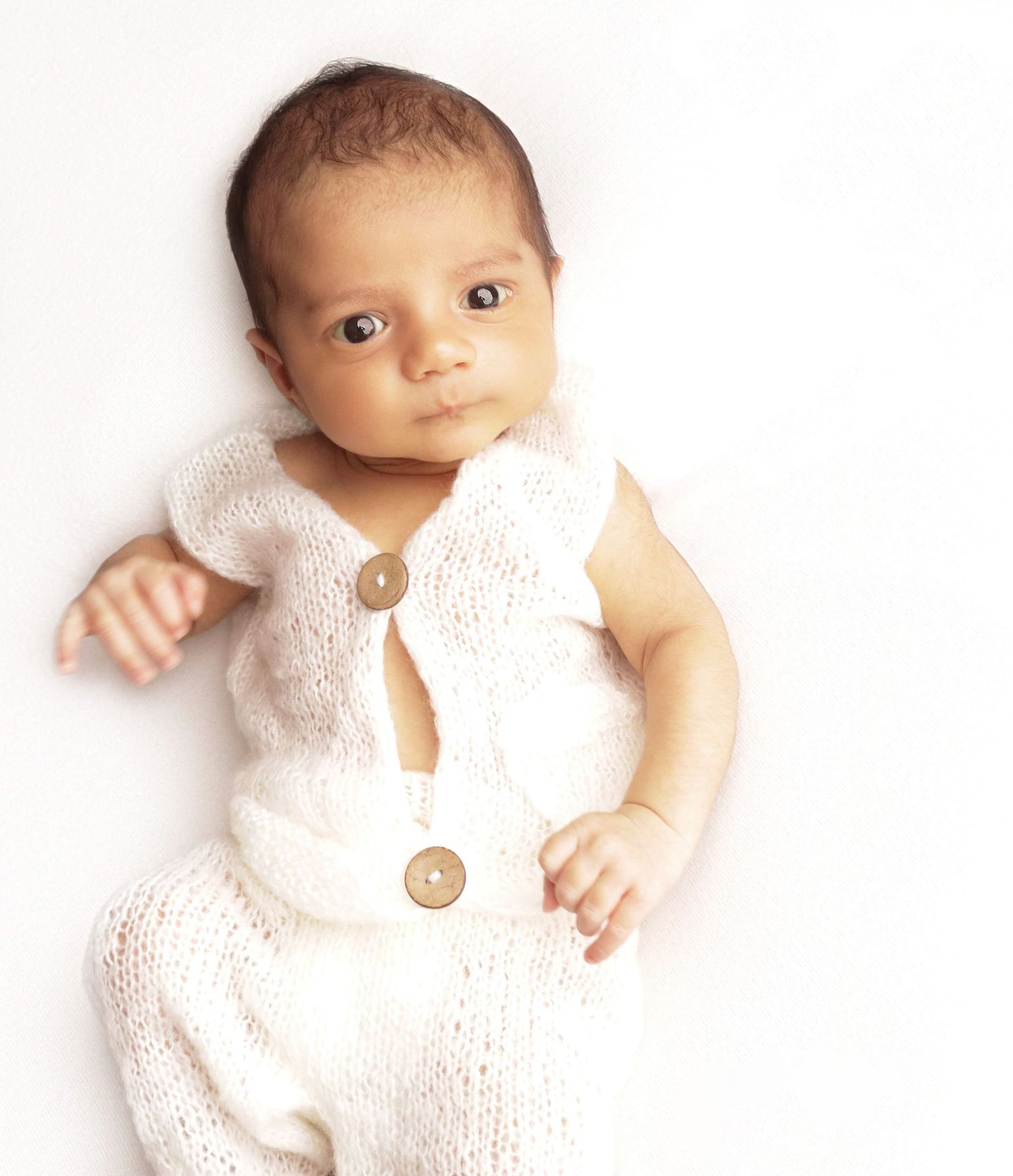 Baby wearing white knit cardigan with buttons lying against light background in studio portrait.