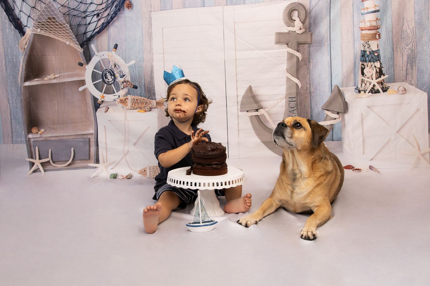 Small child sits next to dog on floor during a cake smash photoshoot with grey and white decorative backdrop.