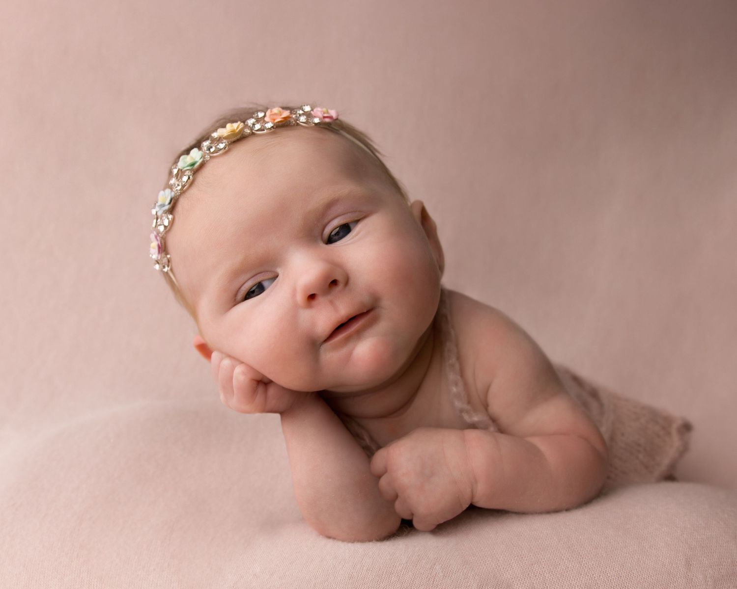 Newborn baby lying on pink blanket wearing delicate floral headband and smiling at camera.