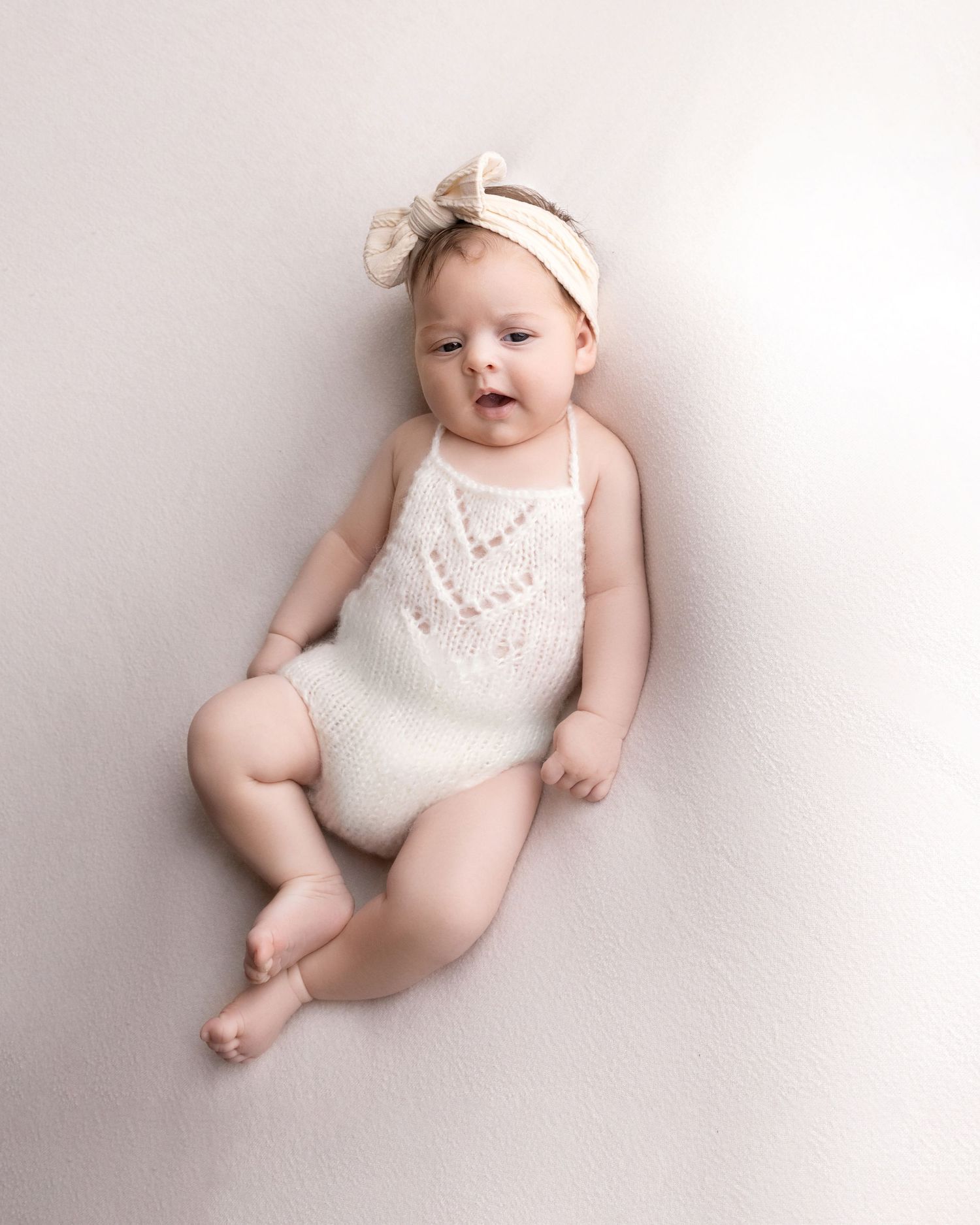 Baby in white lace romper with bow headband posed on light pink background.