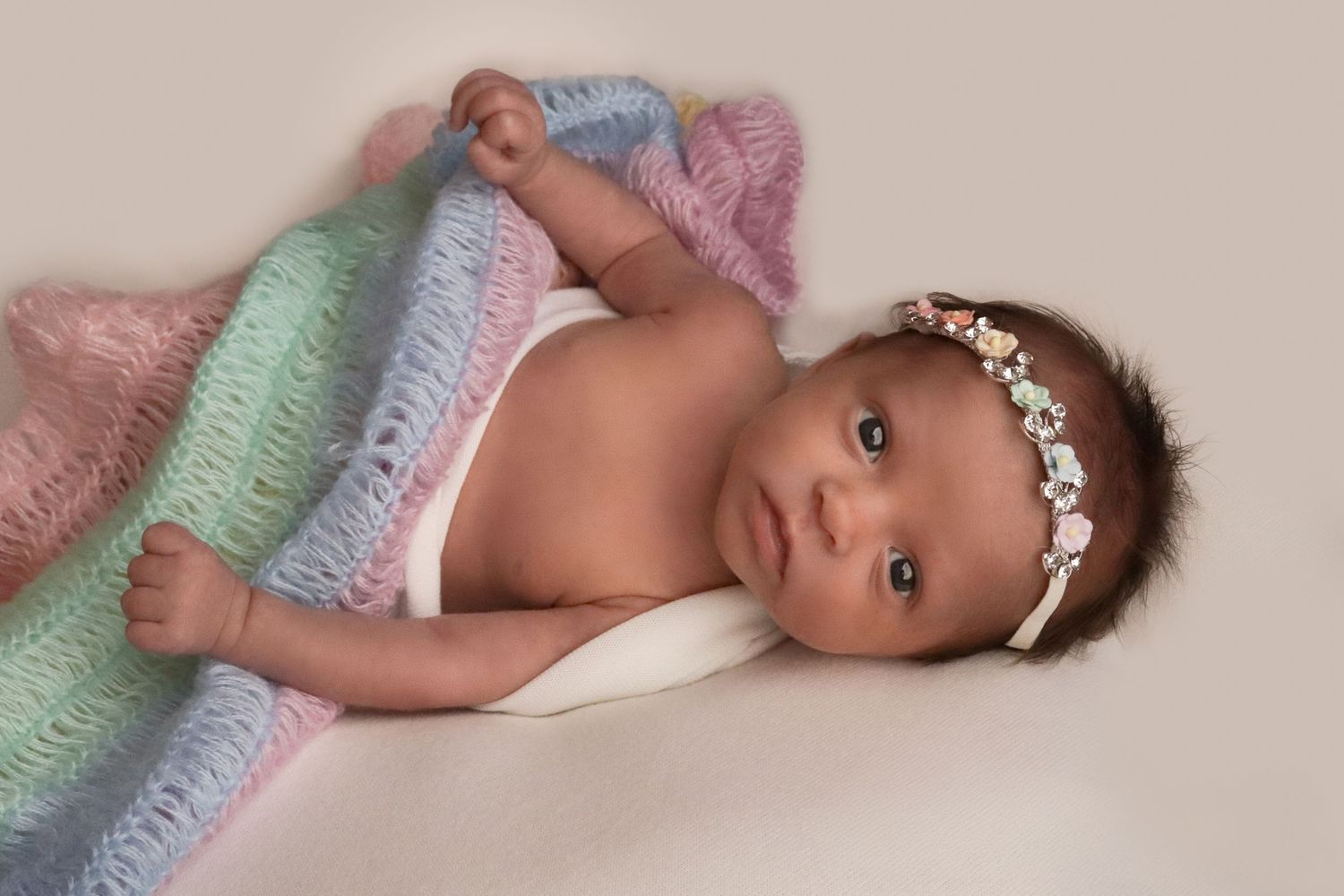Baby lying on soft pastel rainbow blanket wearing flower crown headband in studio.