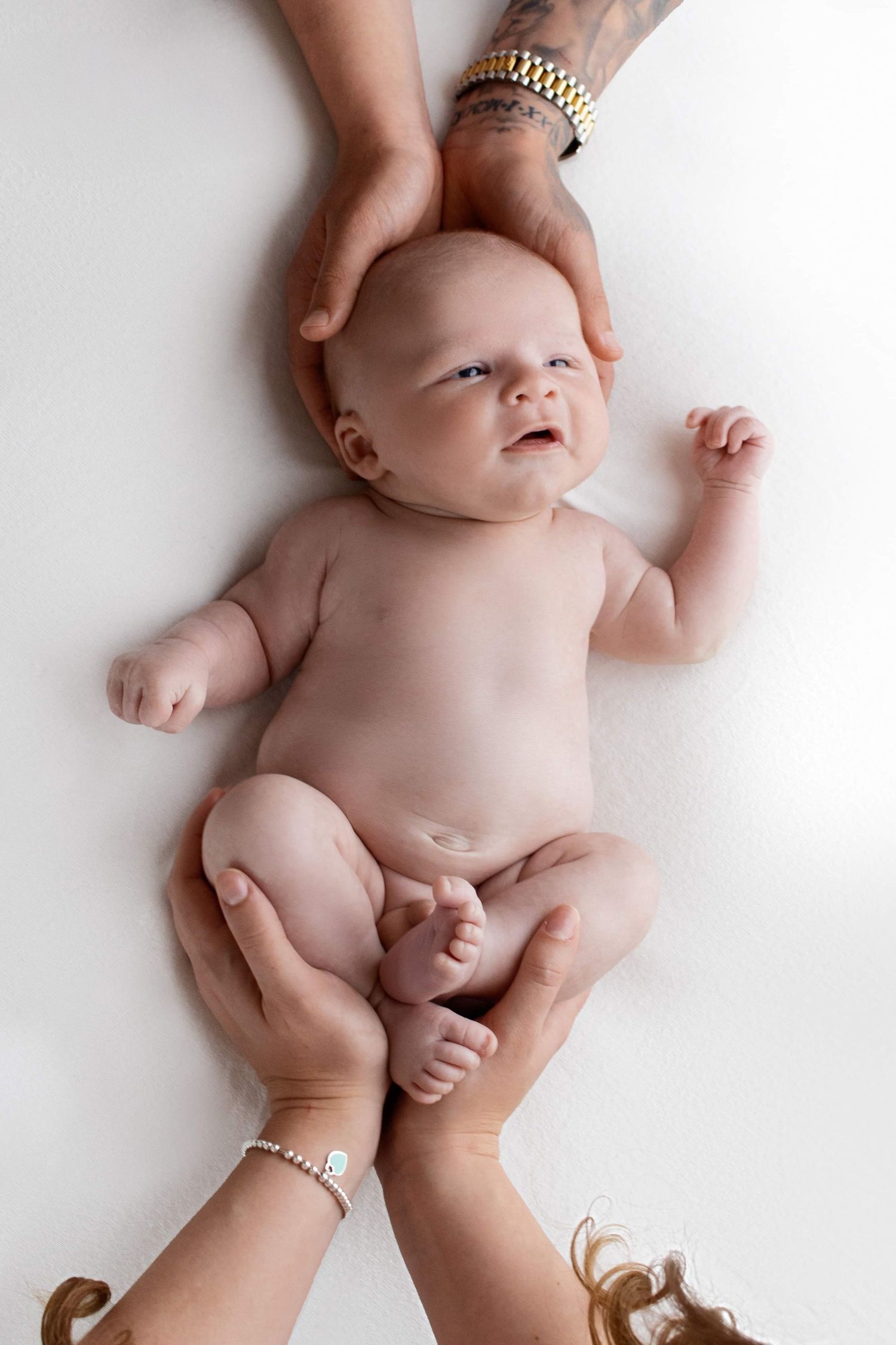 Baby lying on white surface supported by hands wearing jewelry from above.