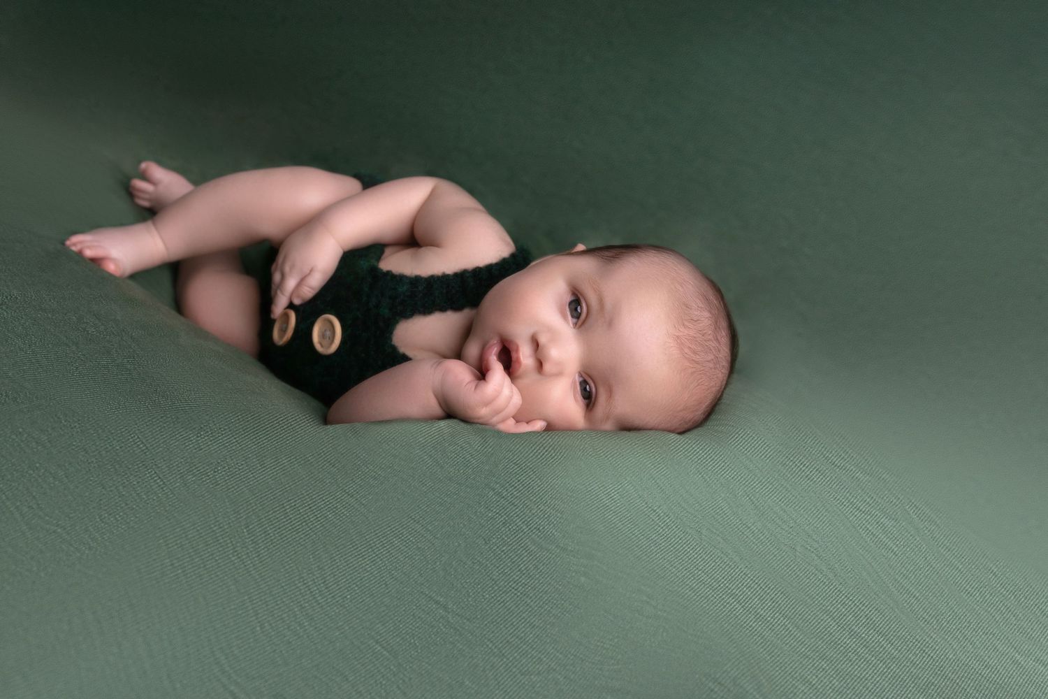 Baby wearing black romper resting sideways on green background during professional photoshoot.