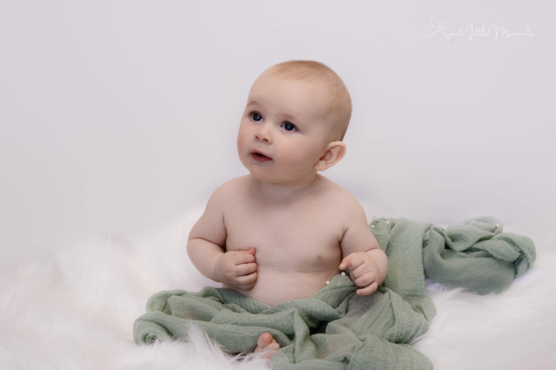 Baby sitting on soft white blanket with sage green fabric wrap, looking off to the side.