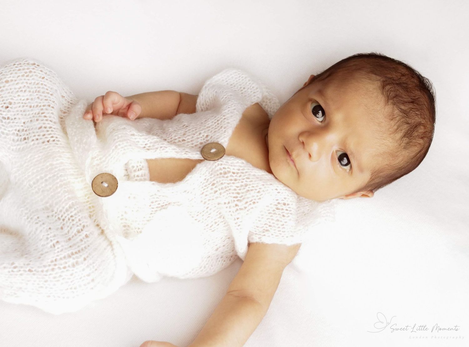 Baby wearing white outfit with decorative buttons looking up at camera.