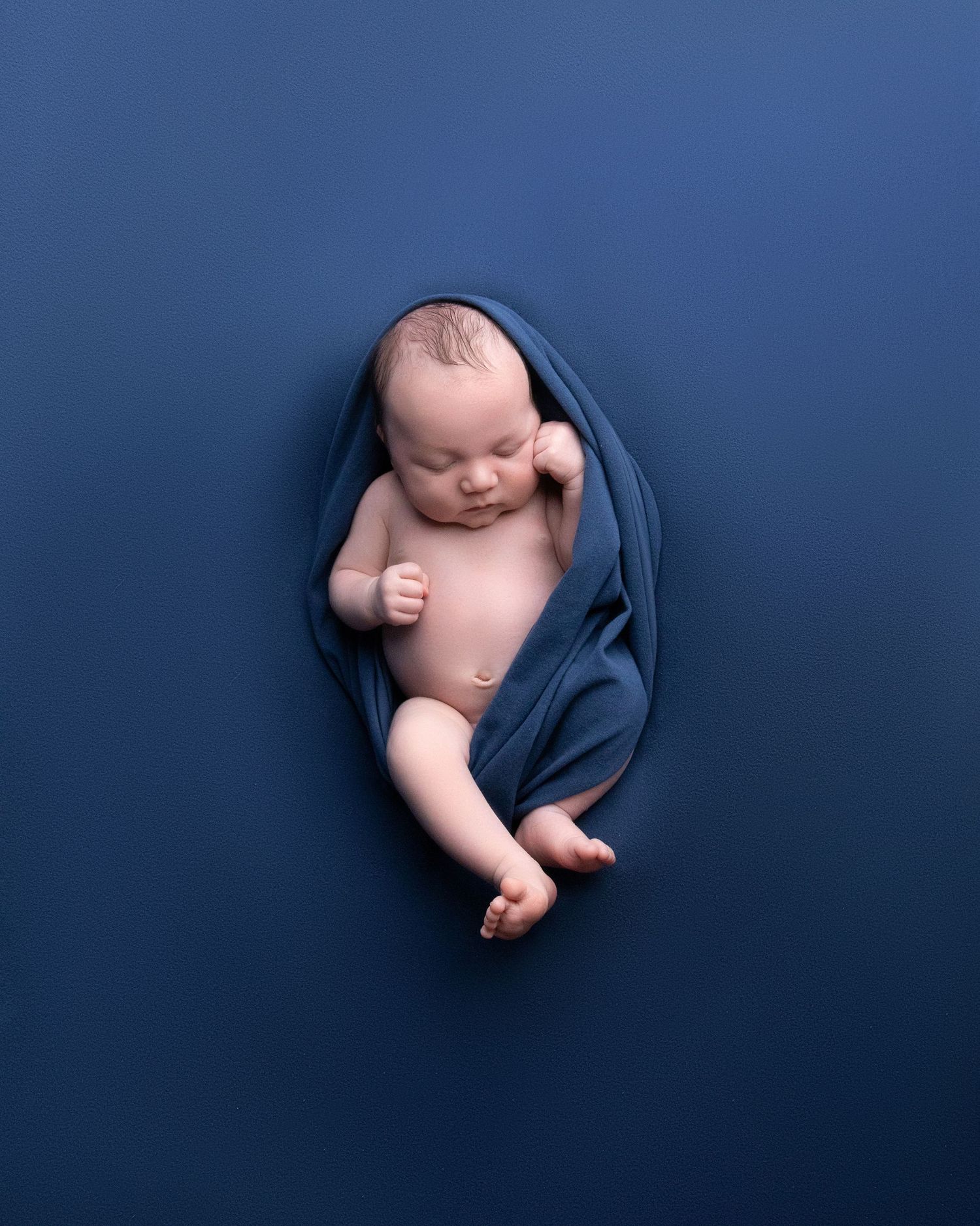 Newborn wrapped in navy blue blanket against dark blue background in minimalist portrait.