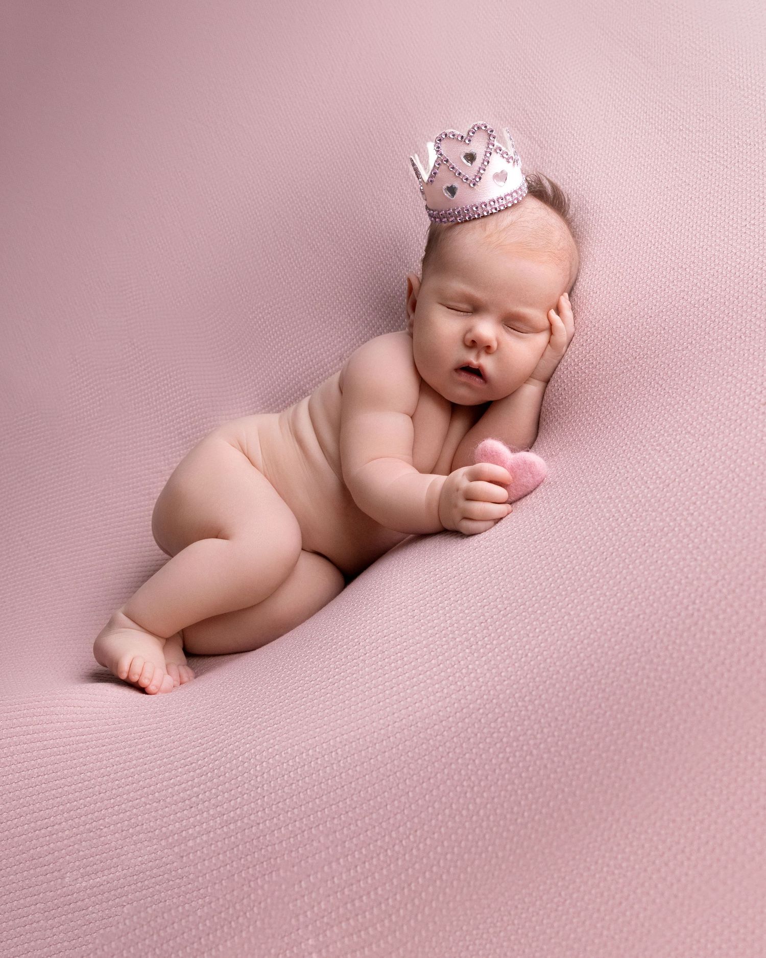 Baby wearing tiny crown sleeping on pink textured backdrop in studio portrait.