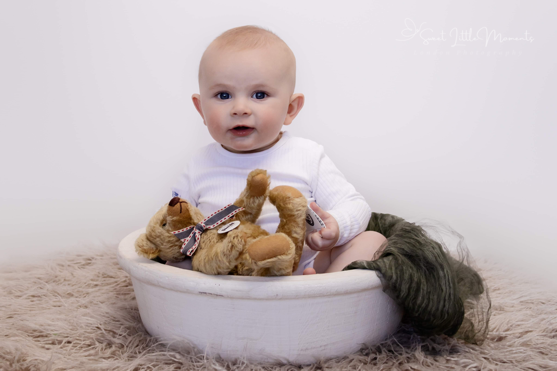 Baby in white shirt sitting with collection of stuffed animals in white bowl.