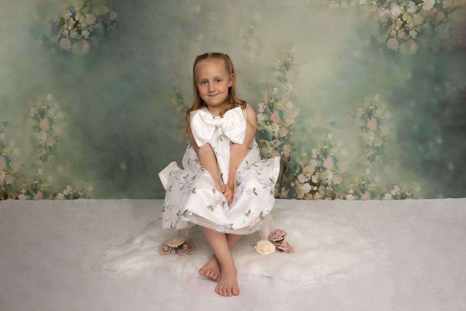 Child in white dress poses against floral backdrop during professional portrait session.