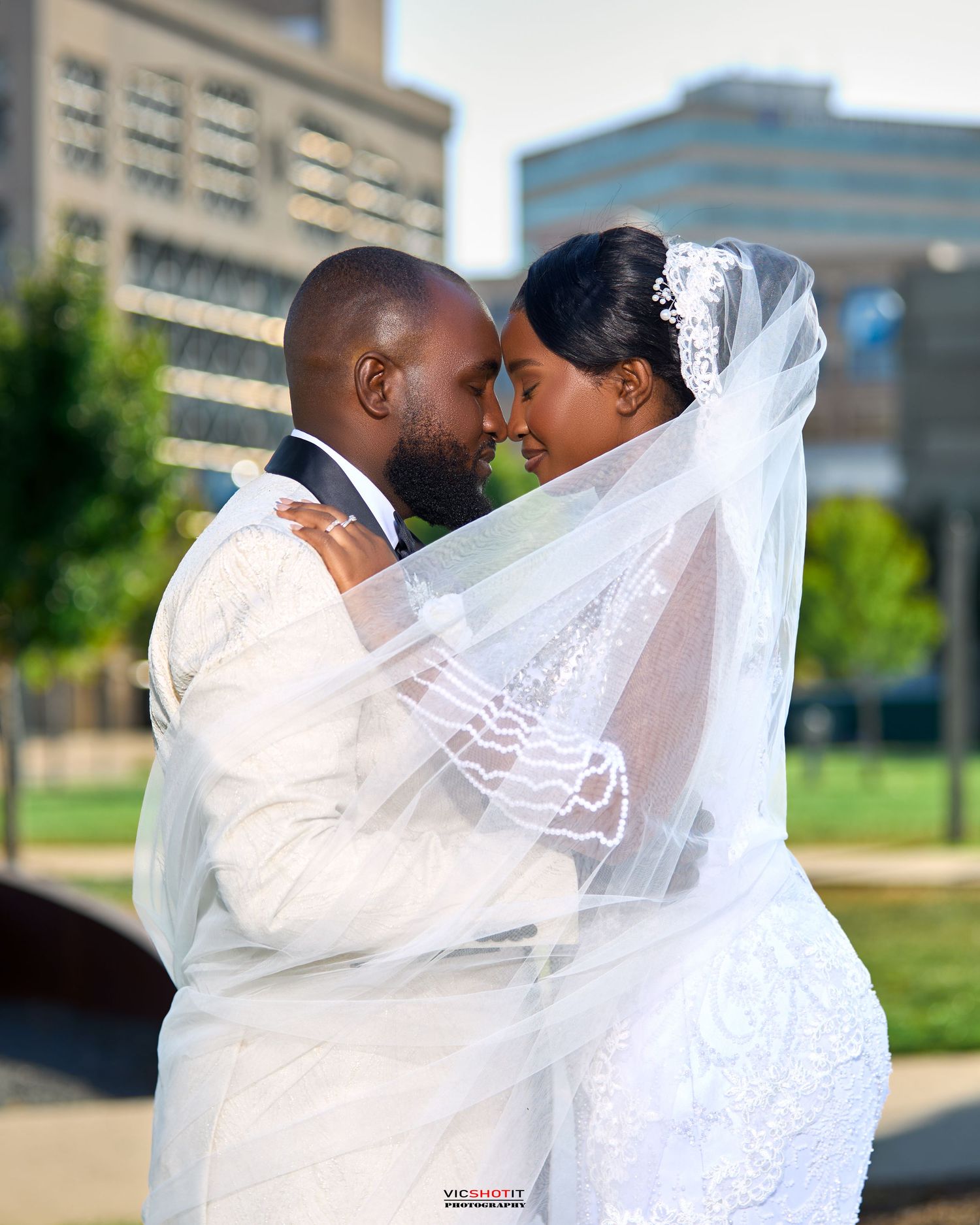 Newlyweds embrace outdoors with flowing veil against urban backdrop.
