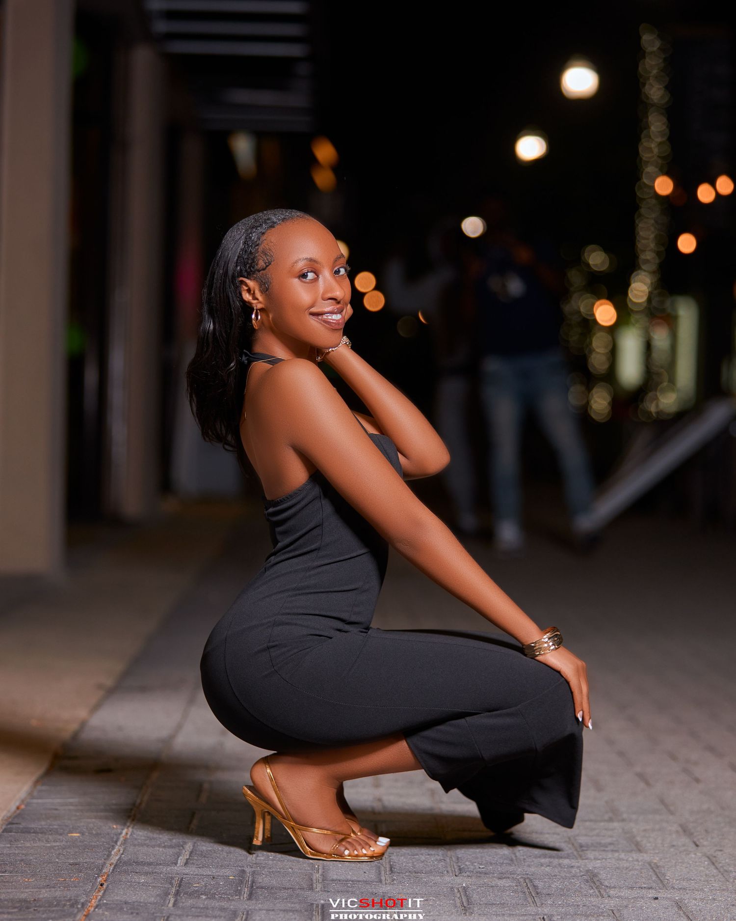An elegant pose in a fitted black dress and gold heels against a nighttime urban backdrop.