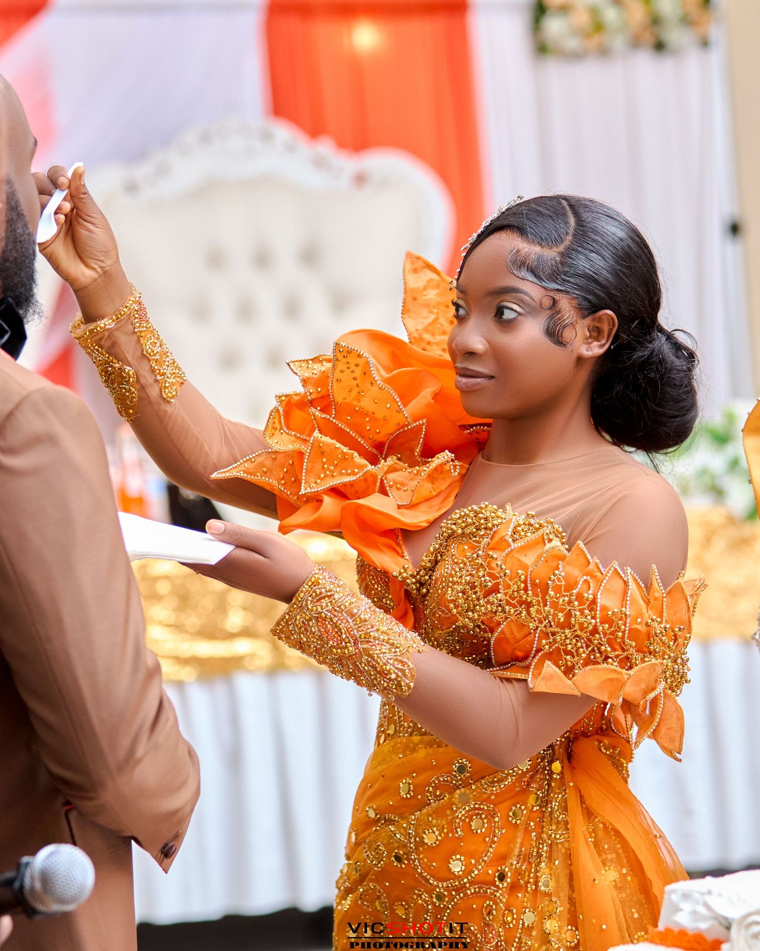 A couple performs a celebratory dance in formal orange and brown attire.