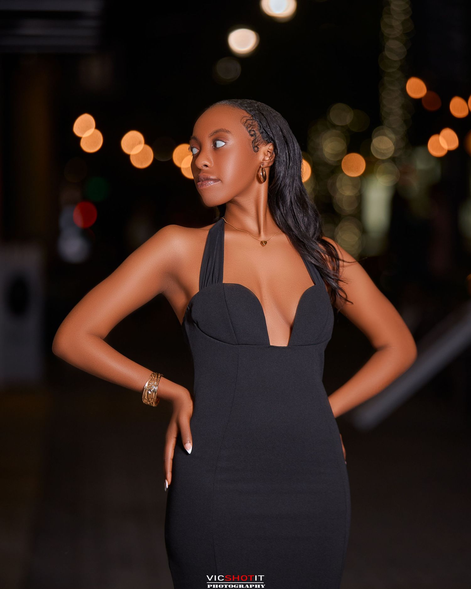 A sleek black formal dress with halter neckline photographed against bokeh lights at night.