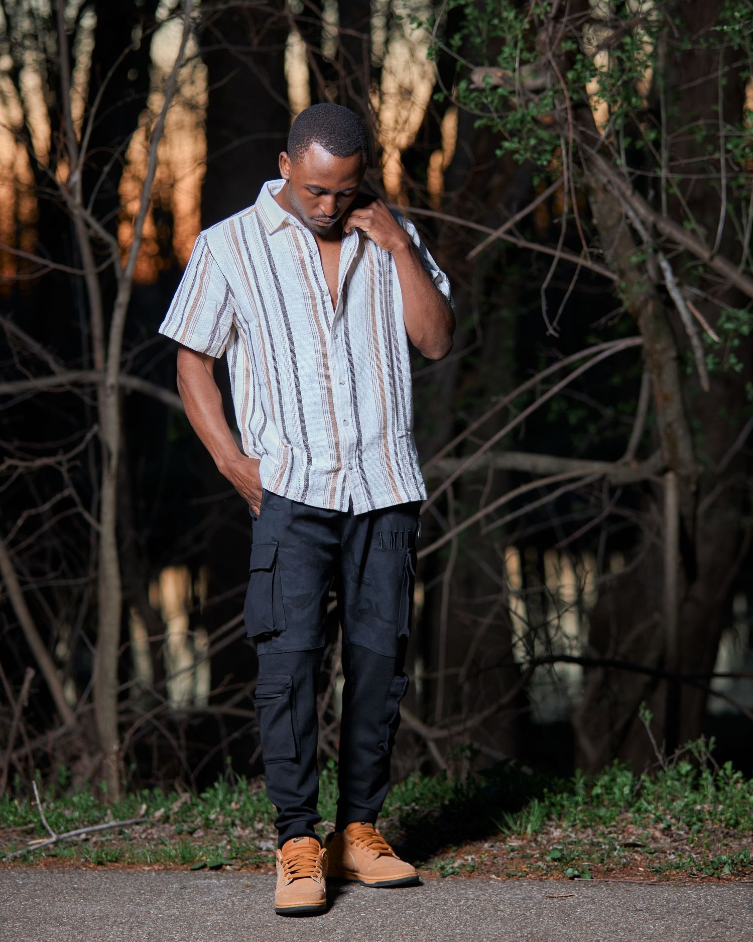 Casual outdoor portrait session showing striped shirt and dark pants against wooded background.