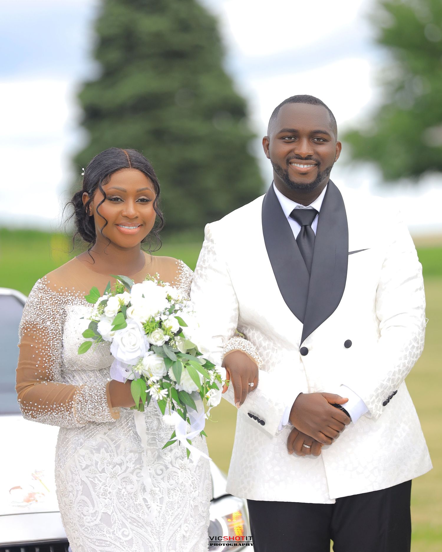 Wedding couple in white and black formal attire posing outdoors with natural backdrop.
