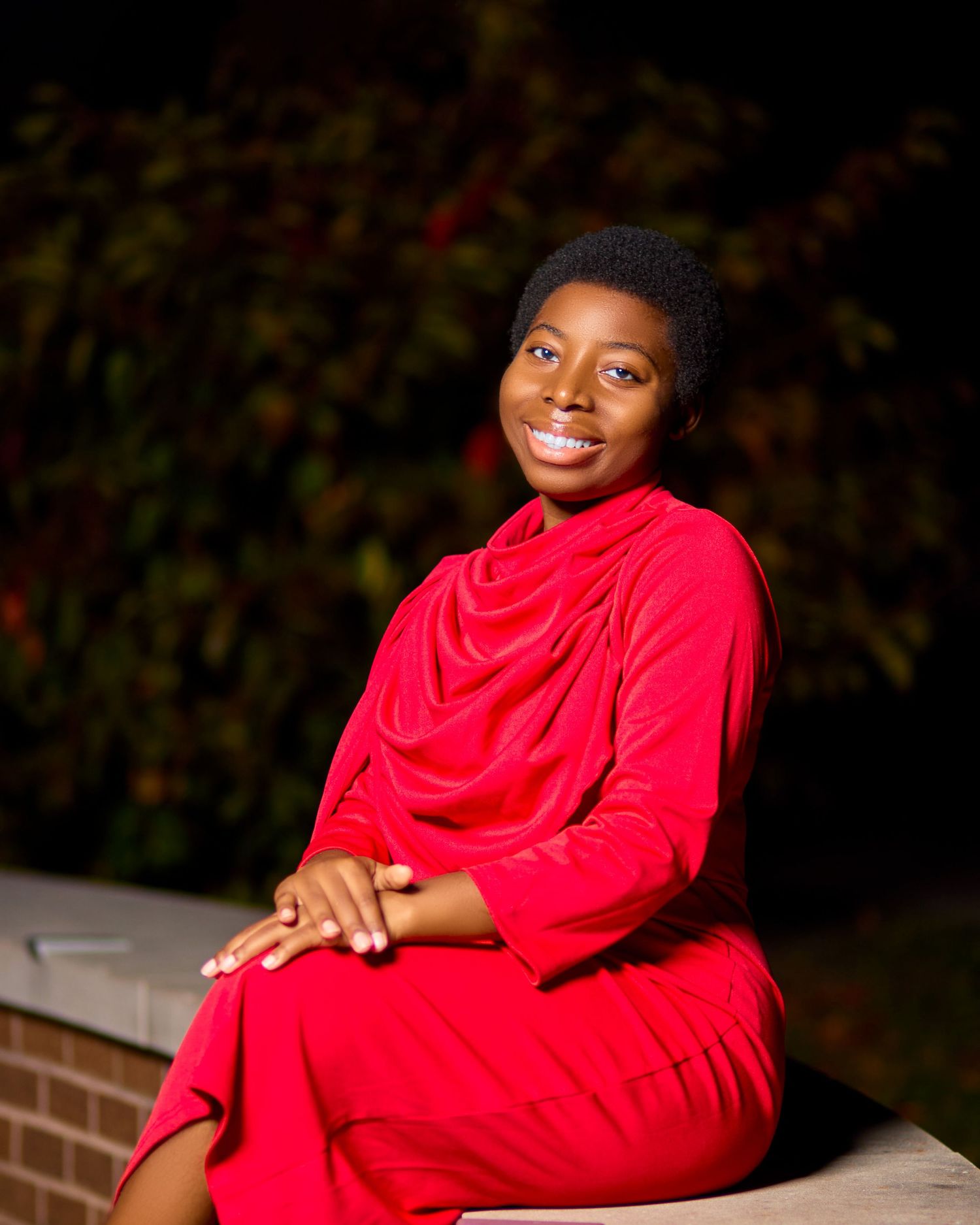 Portrait of a model in a striking red draped dress sitting on brick wall during sunset.