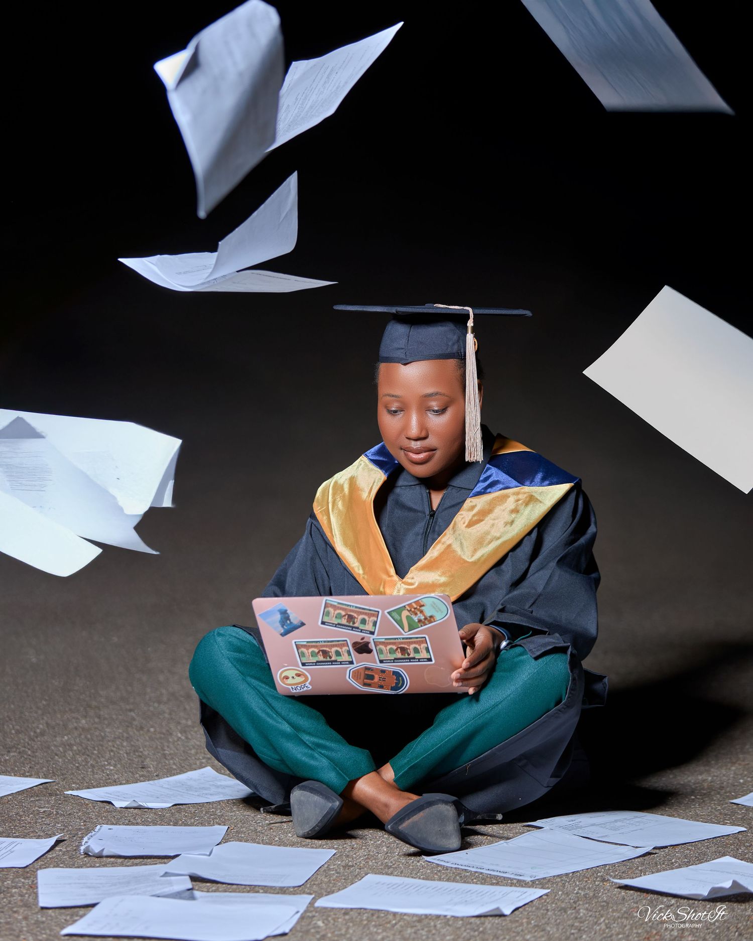 Graduate in cap and gown examines papers scattered on floor in artistic sequence.