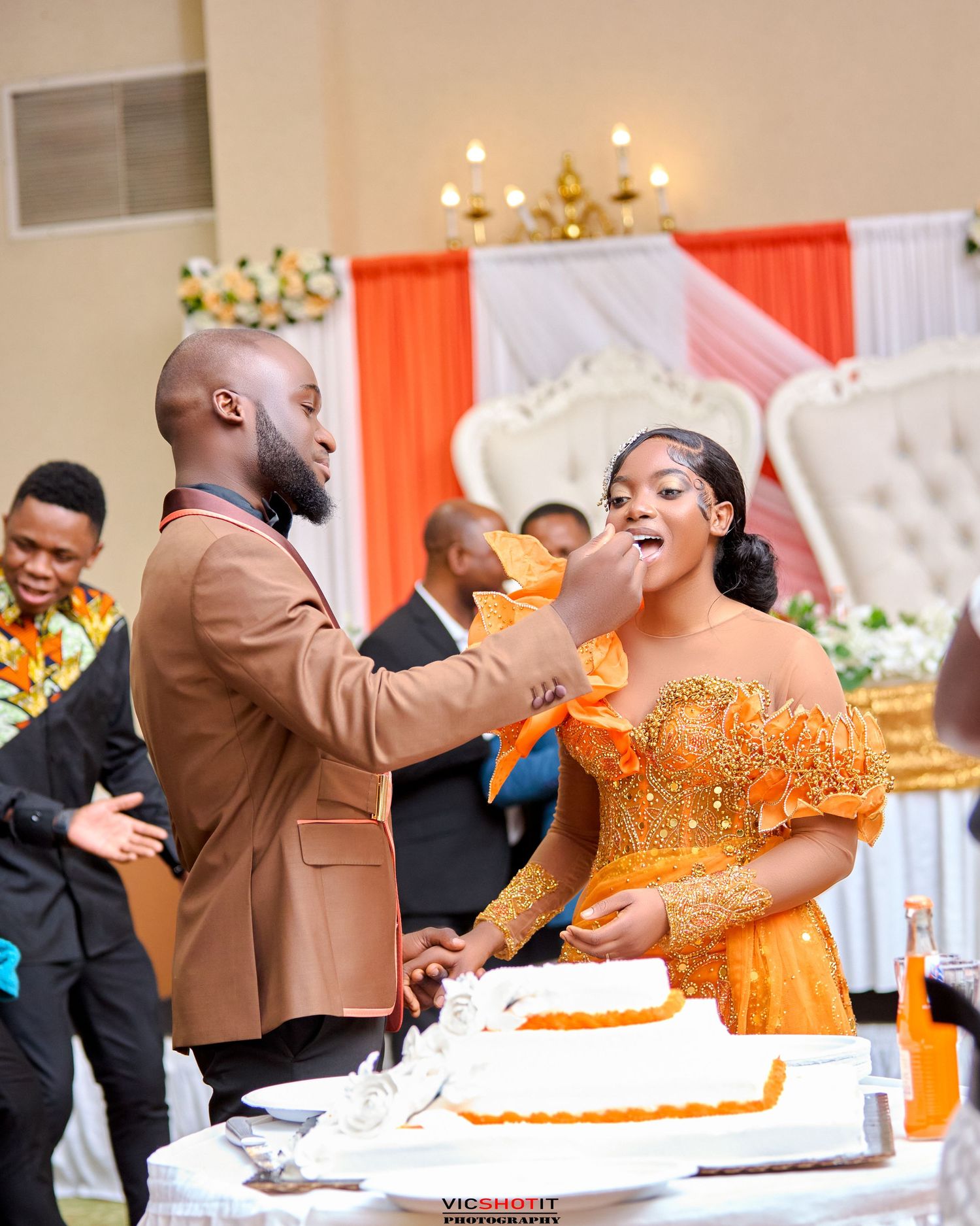Wedding guests celebrate during a lively cake-cutting ceremony at an indoor reception.