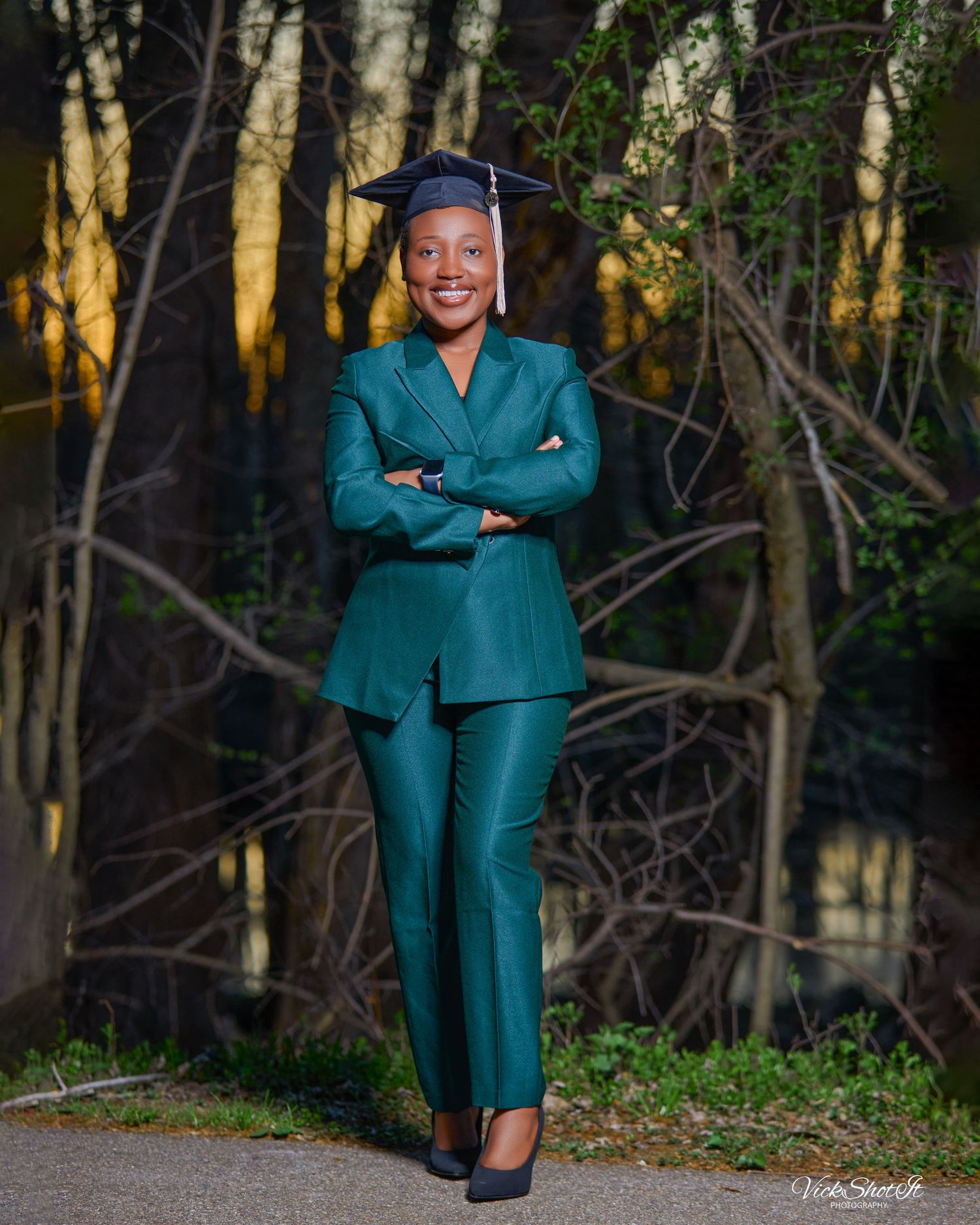 Graduate in green attire poses outdoors against natural forest backdrop.