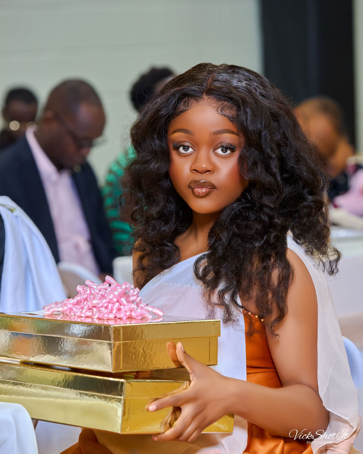 A woman in an orange dress sits with gold gift boxes at a celebration.