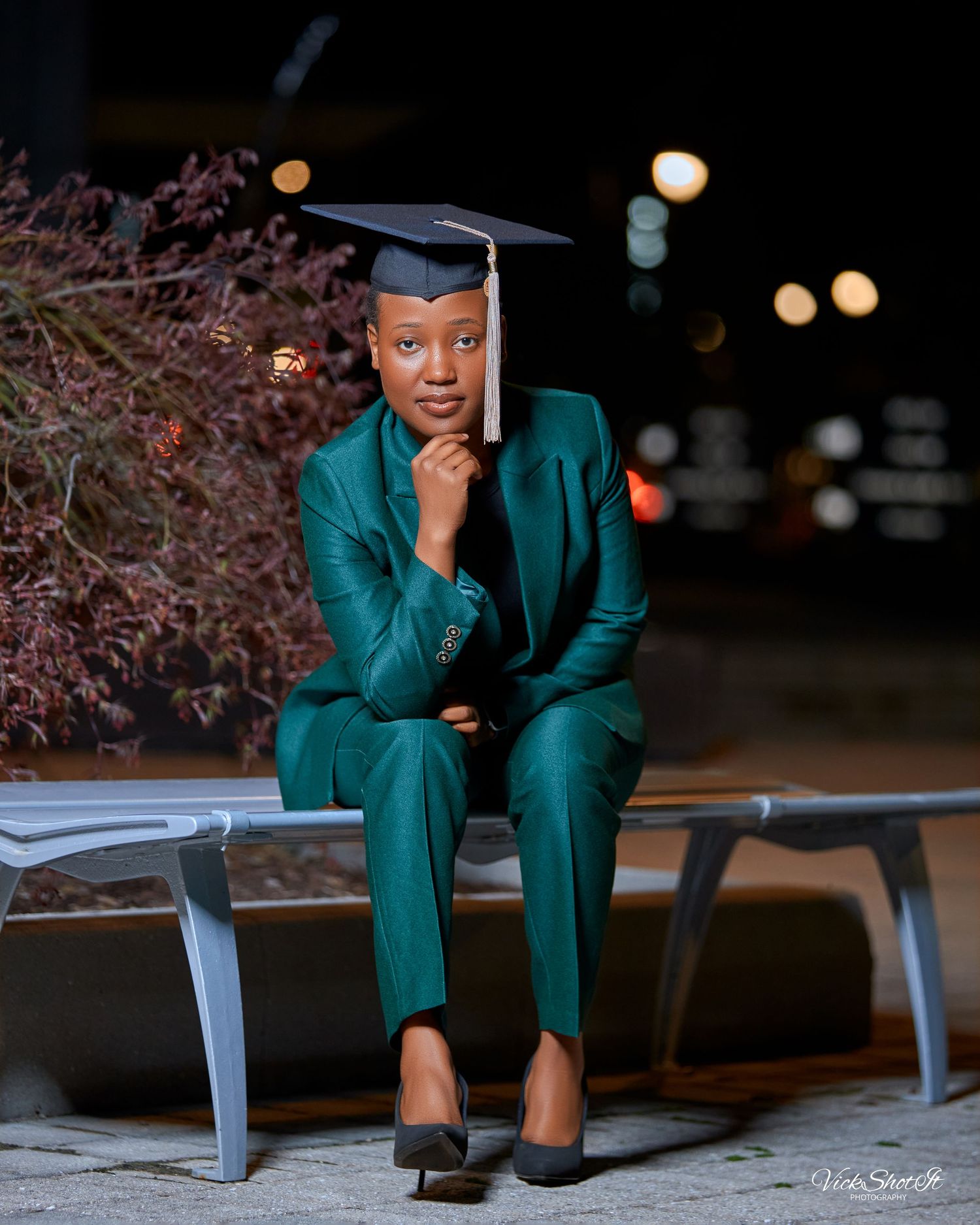 A graduation portrait in a teal business suit with mortarboard cap against evening lights.