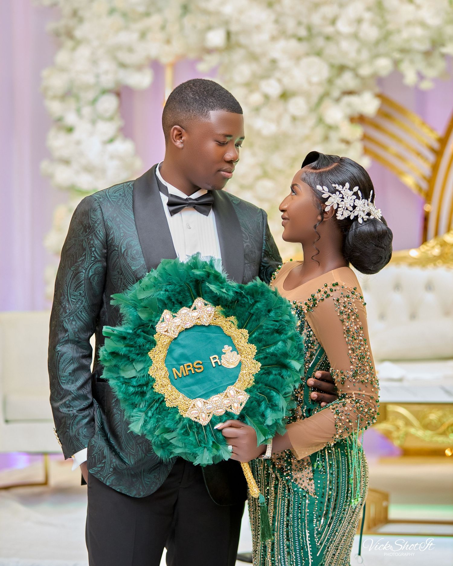 A couple in formal attire posing with an emerald green decorative wreath at a wedding reception.