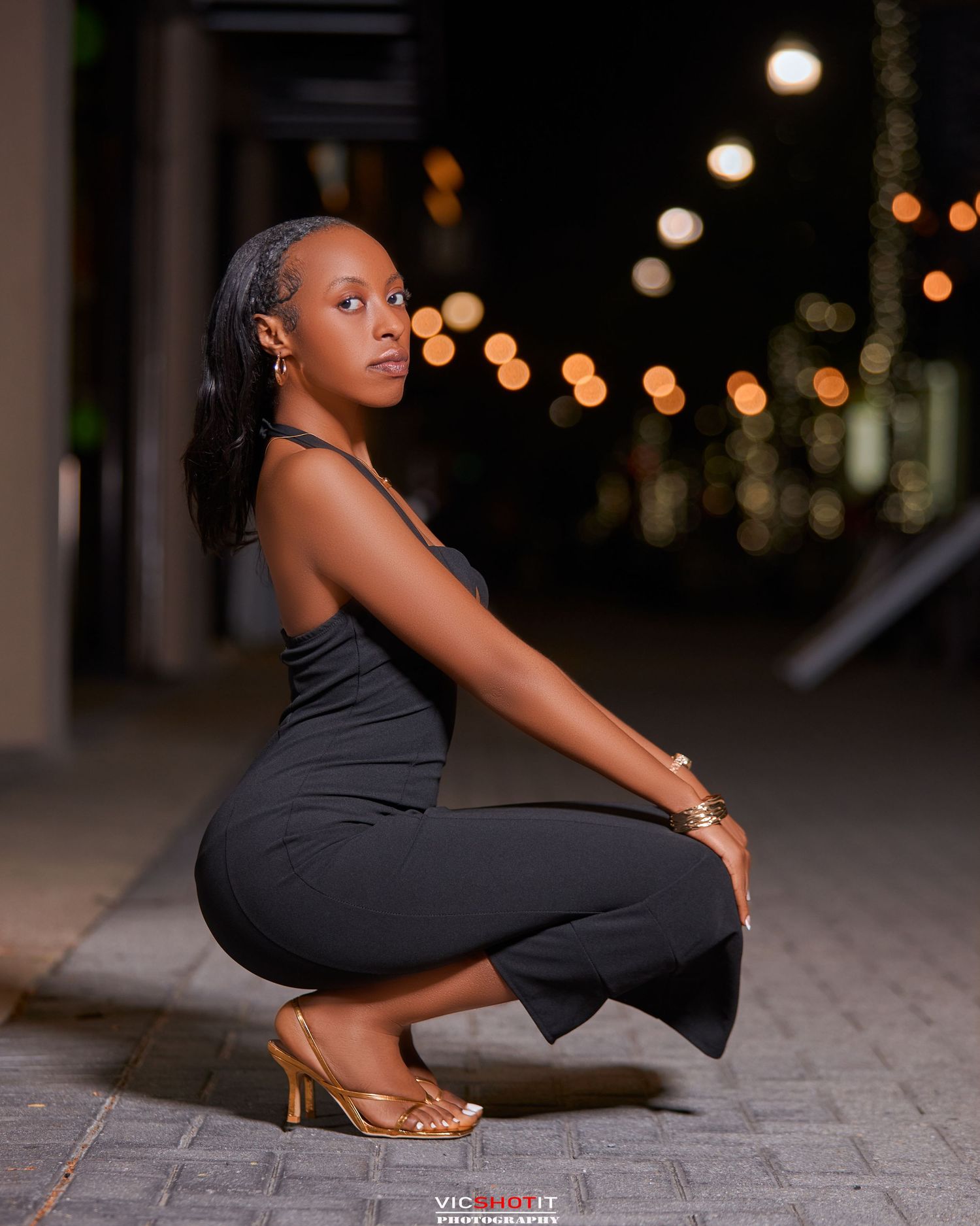 A graceful crouching pose in a black fitted dress captured at night with bokeh lighting.