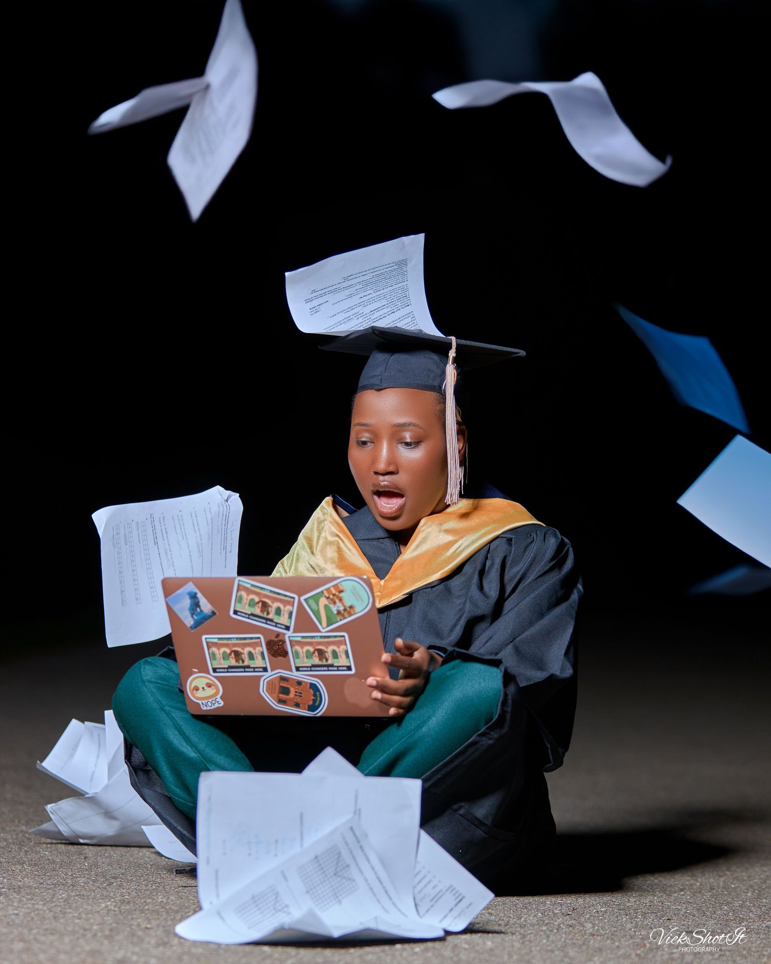 Graduate in academic regalia sits among scattered documents in dramatic lighting.