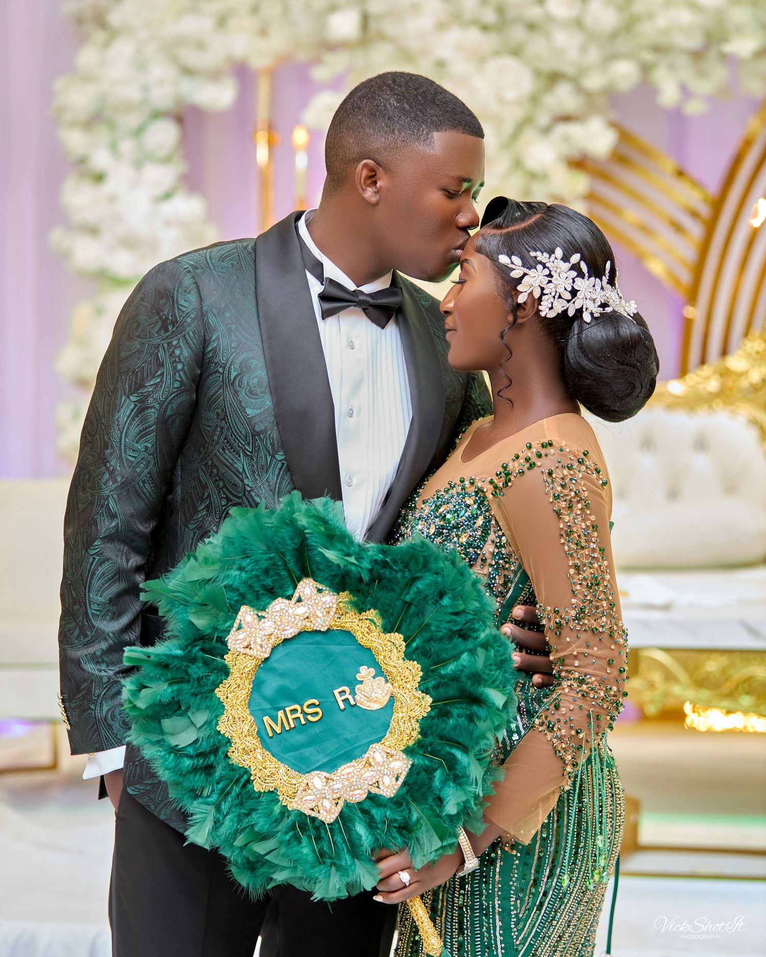 A couple sharing an intimate moment with a green and gold decorative wreath at an elegant event.