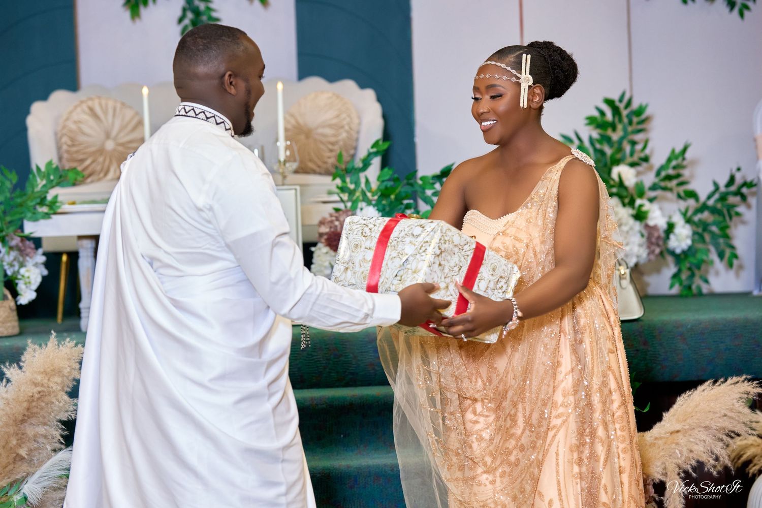 A couple in elegant attire exchanges a gift during what appears to be a wedding ceremony with floral decorations.