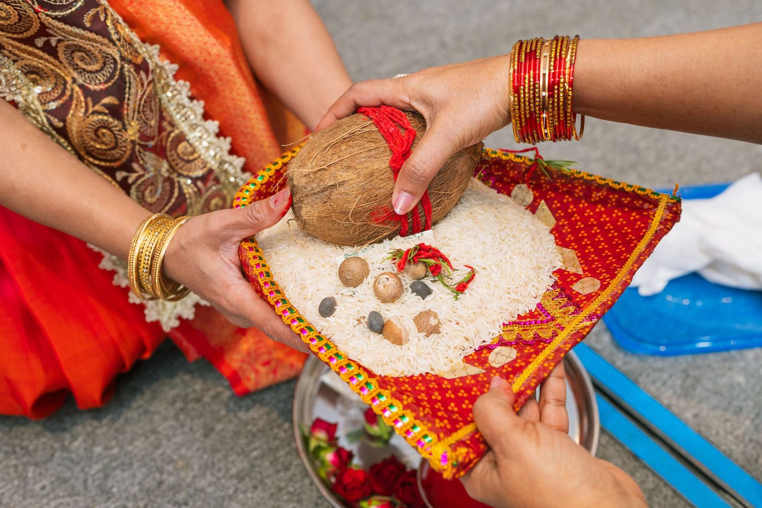 Hands adorned with bangles and jewelry arrange coconut and offerings on a decorative red cloth for a traditional ceremony.