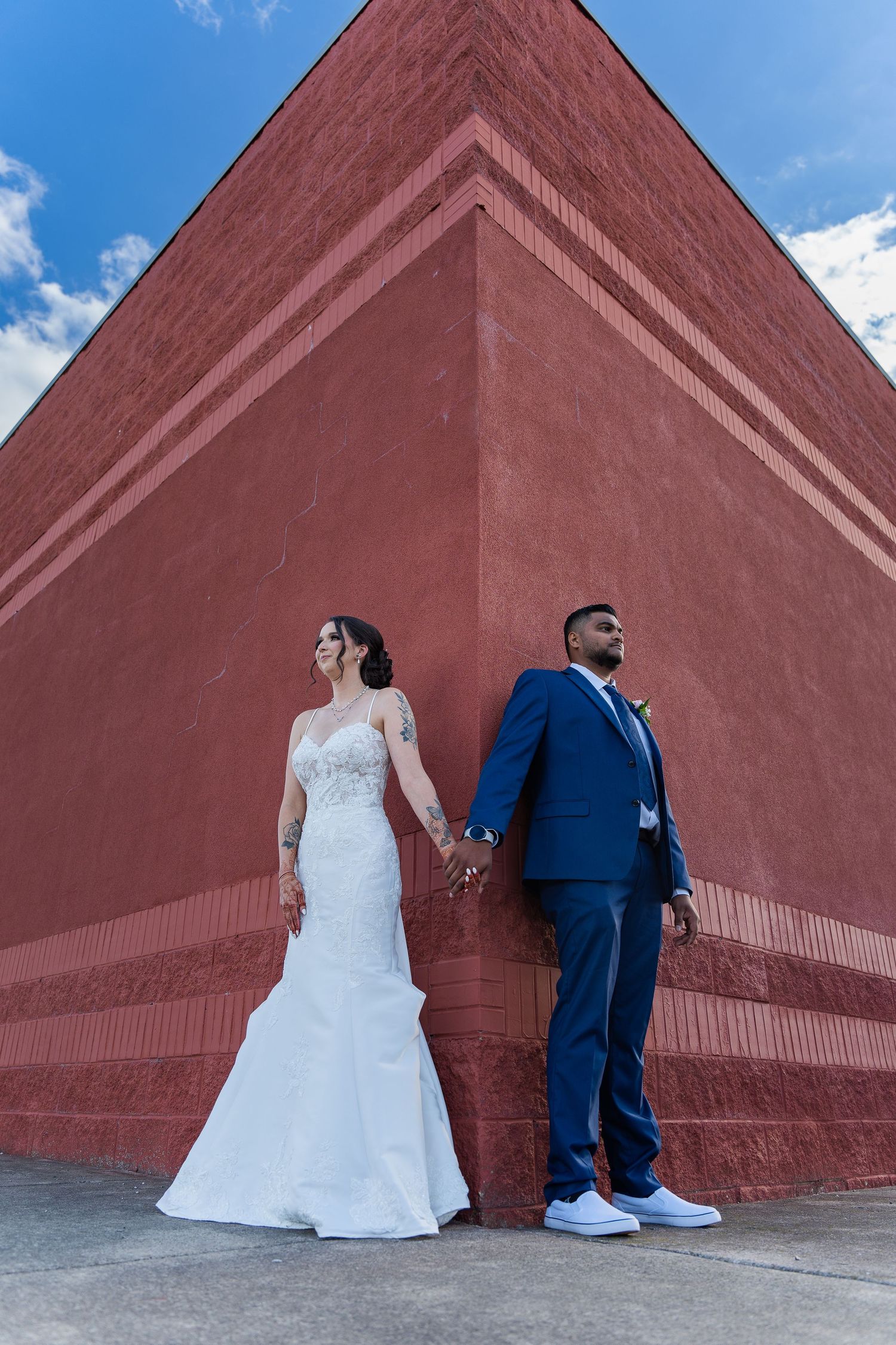 Wedding couple poses dramatically against a large terracotta wall while holding hands.