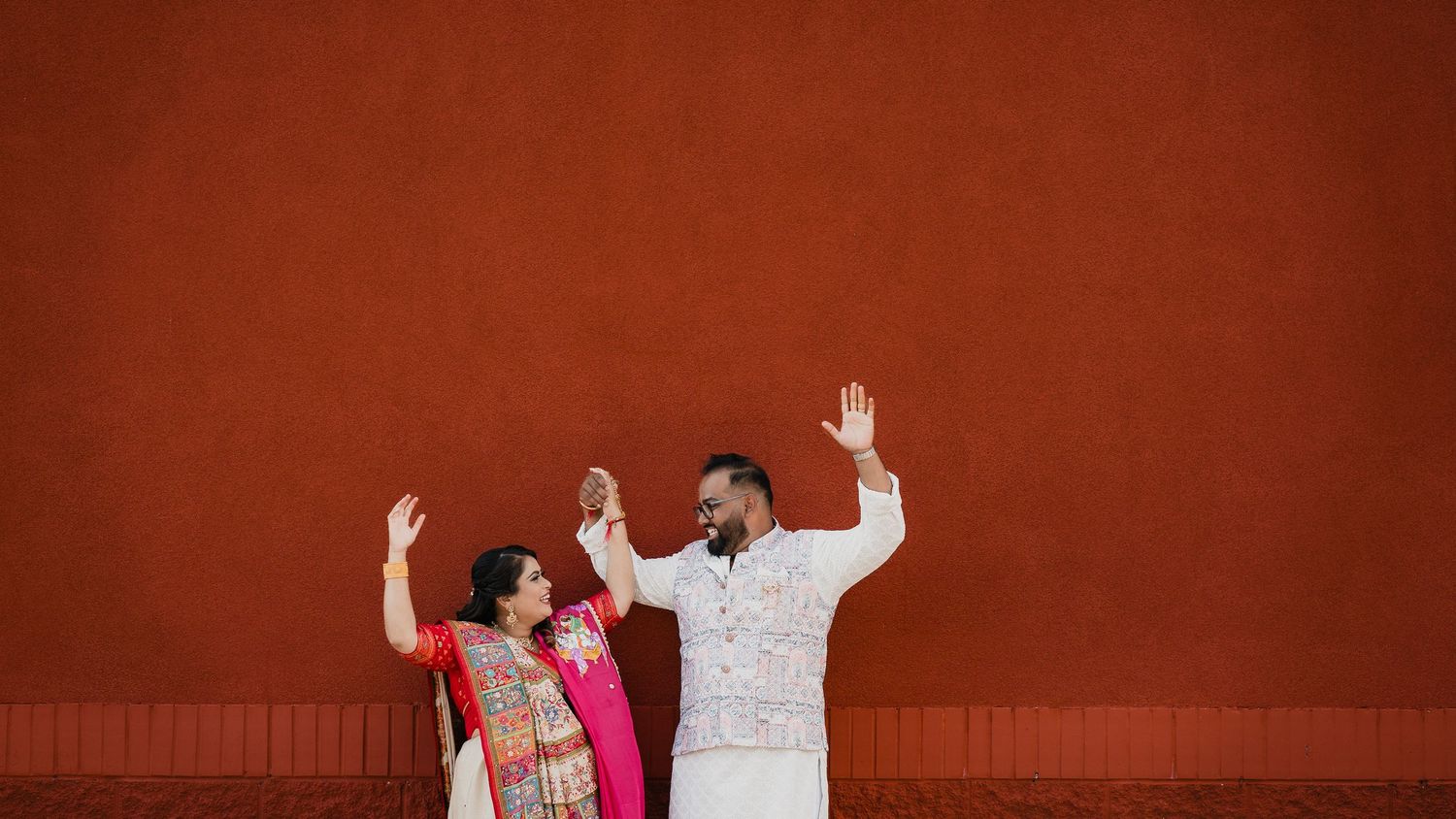Three people in traditional Indian clothing pose joyfully against a bright red wall background.