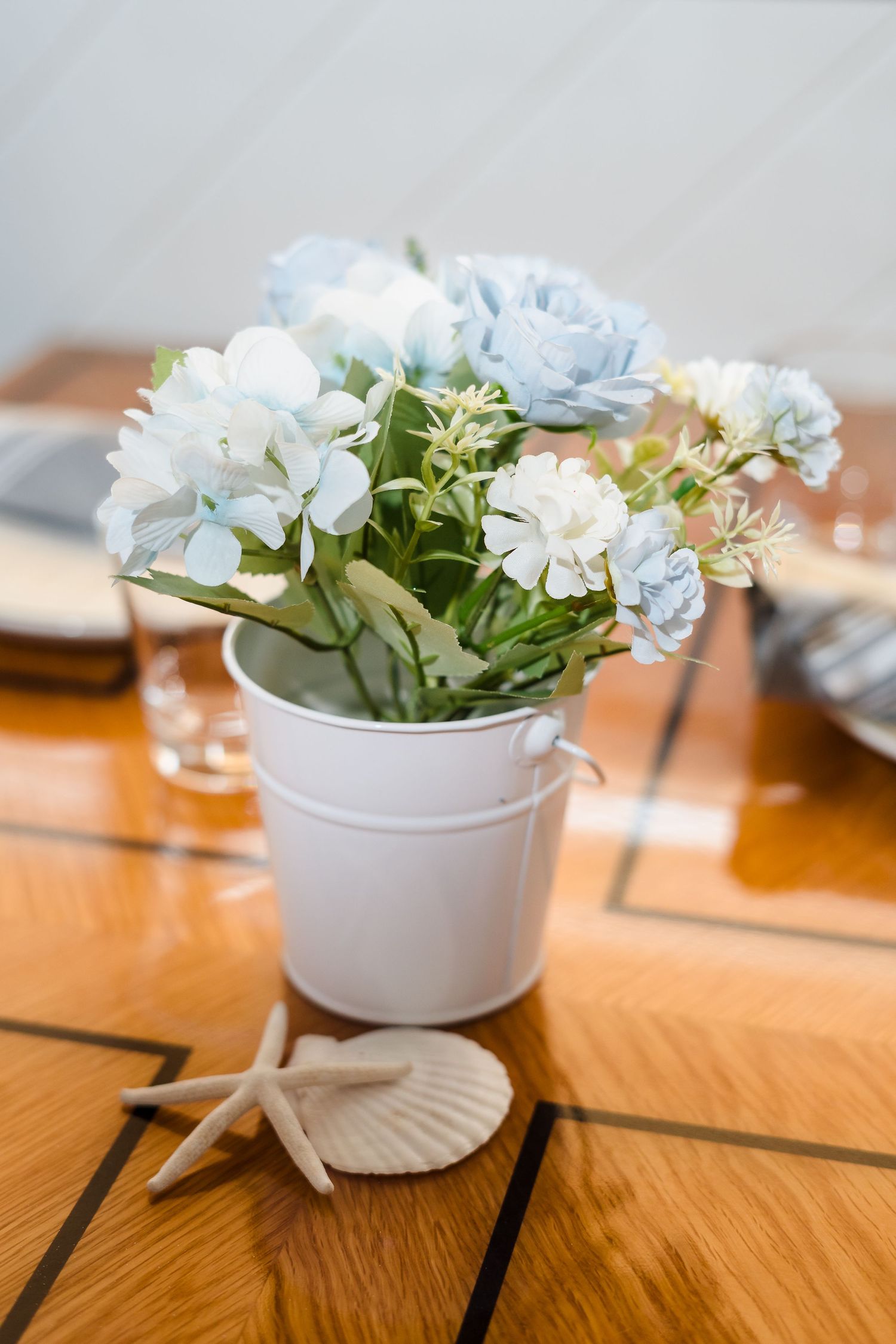 Small white bucket vase filled with light blue and white flowers sits on wooden table with decorative starfish.