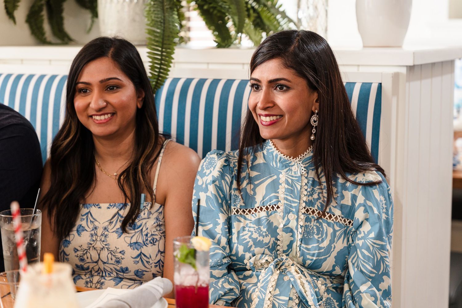 Two friends in blue and silver outfits share a joyful moment at a formal celebration with drinks and decor.