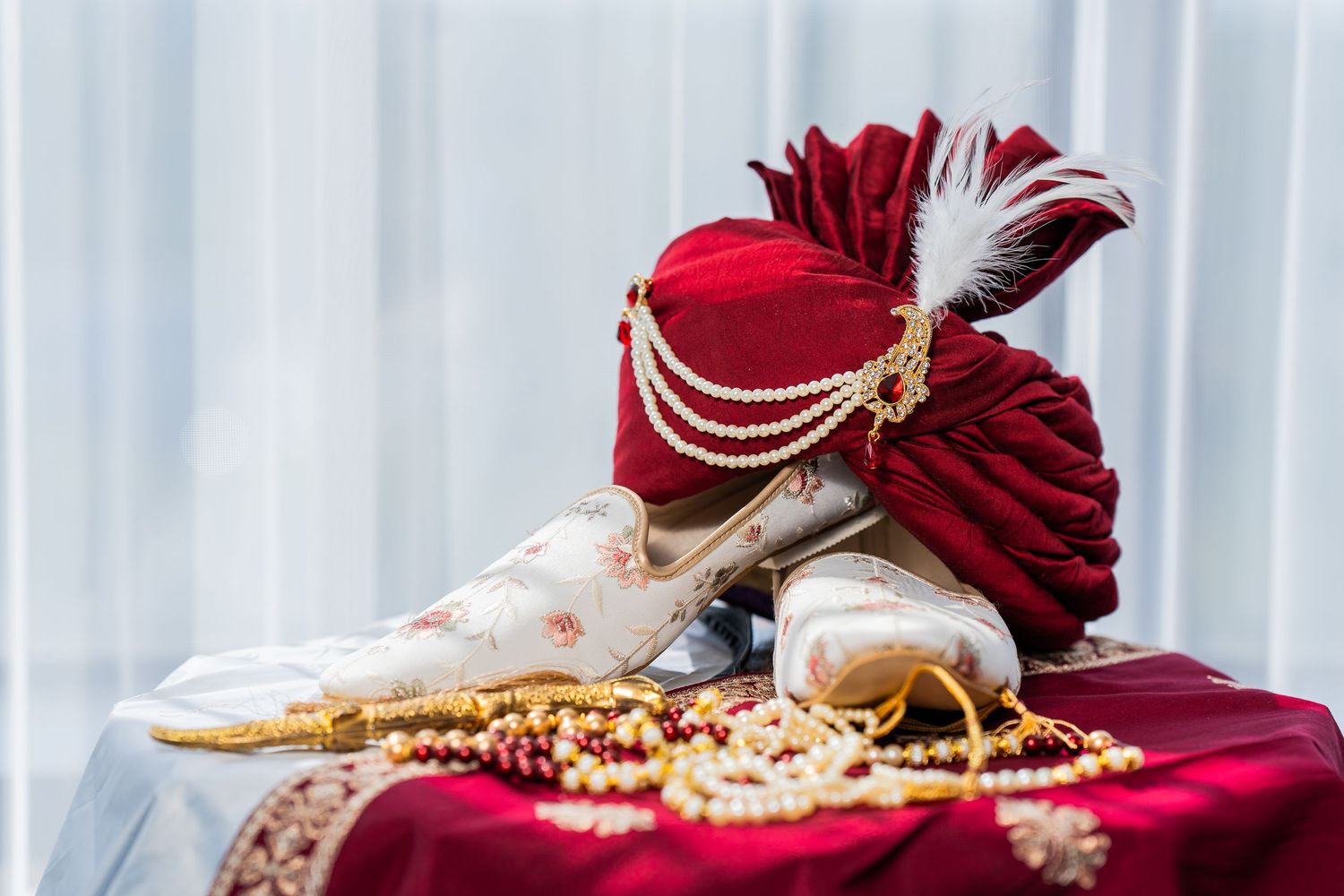 Traditional Indian groom accessories including red turban with pearls and shoes on burgundy fabric.
