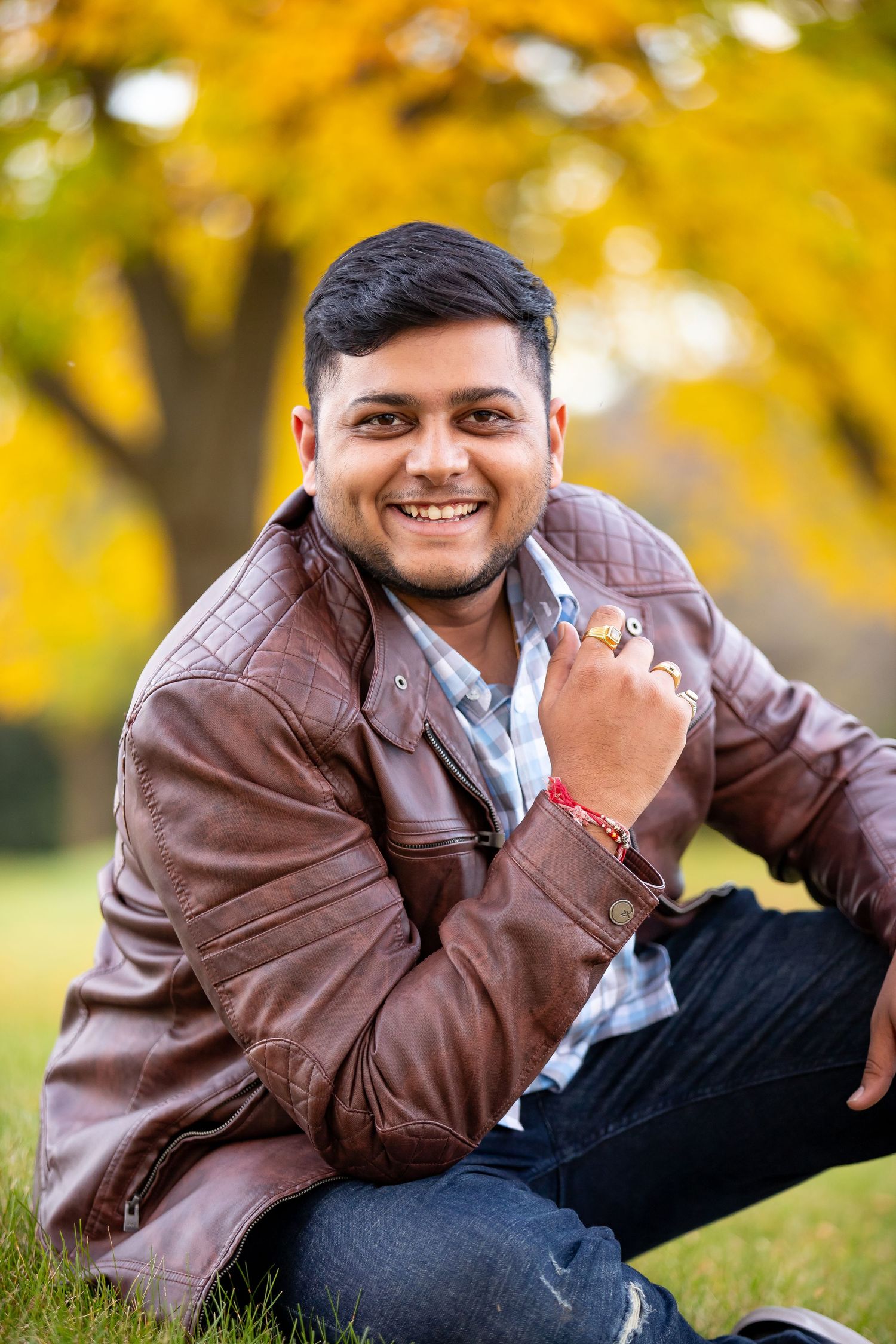 Smiling young person in brown leather jacket sitting on grass with autumn foliage in background.