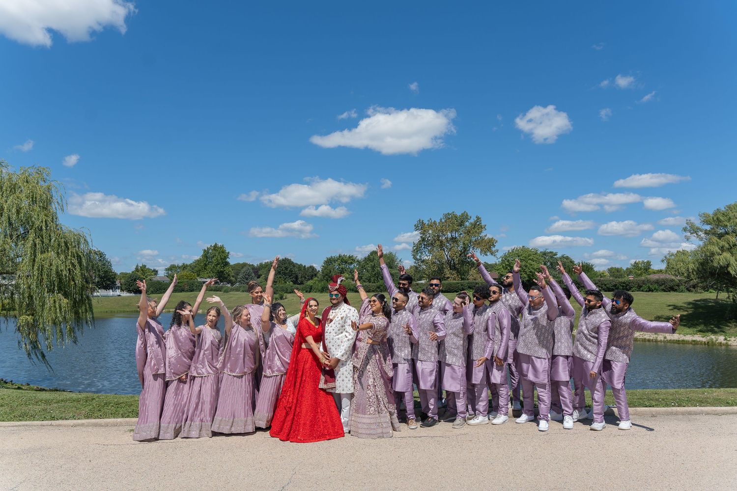 Wedding party in purple attire poses with couple by lake under blue sky.