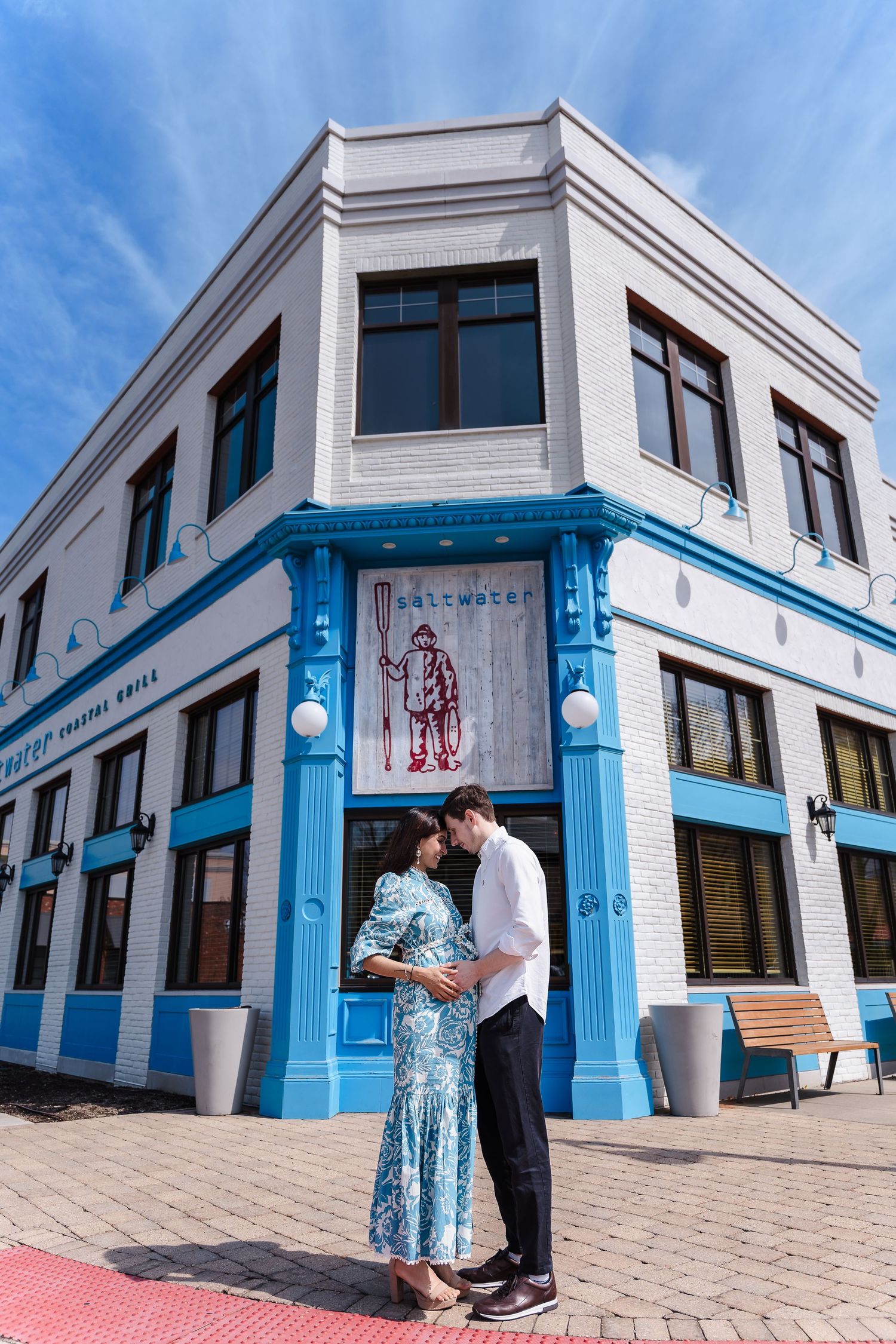 A couple embraces outside a white and blue corner building on a sunny day with decorative architectural details.
