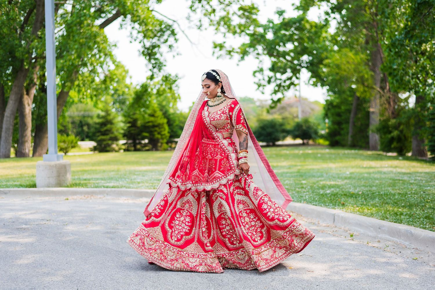 Bride in stunning red lehenga with intricate embroidery poses in a lush green park setting.