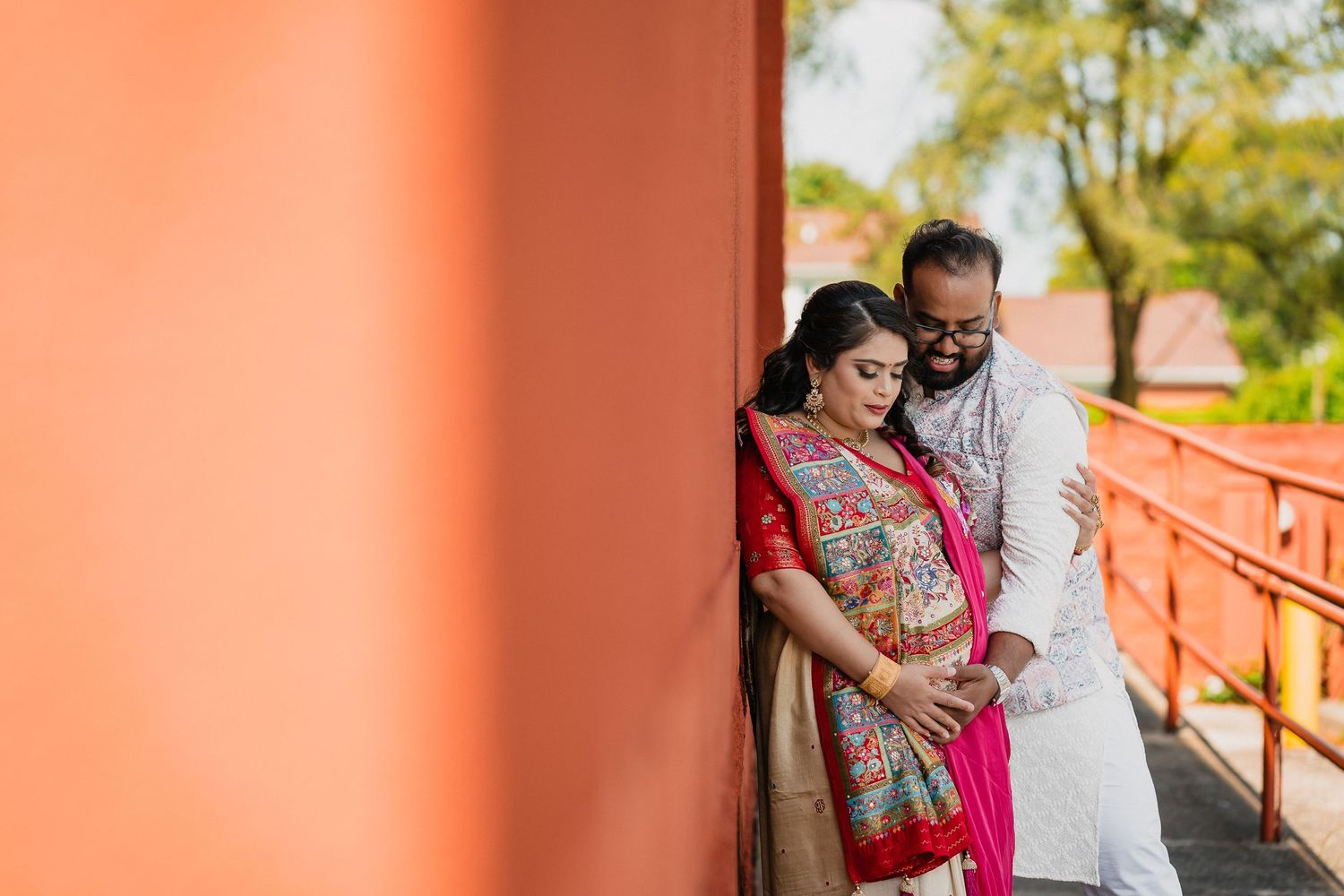 A couple in traditional Indian attire pose together against a coral-colored wall during a maternity photoshoot.